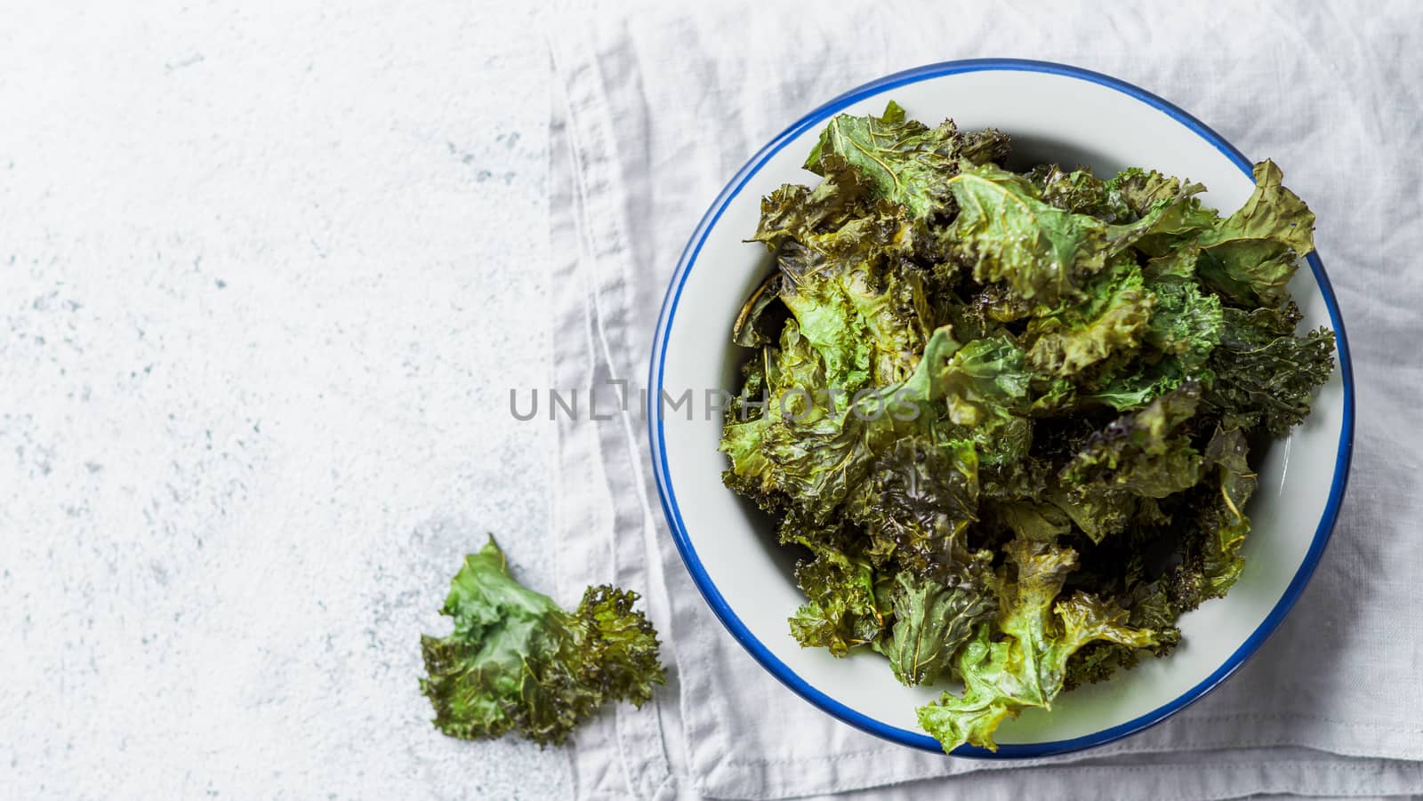 Green Kale Chips with salt on plate. Homemade healthy snack for low carb, keto, low calorie diet. Gray cement background. Ready-to-eat kale chips, copy space for text. Top view or flat lay. Banner