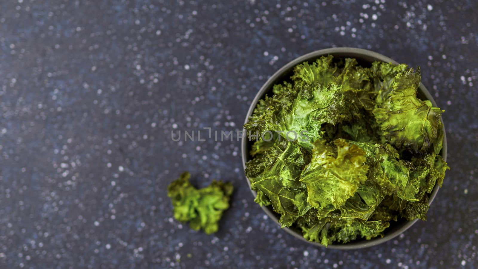 Green Kale Chips with salt in bowl. Homemade healthy snack for low carb, keto, low calorie diet. Dark blue background. Ready-to-eat kale chips, copy space for text. Top view or flat lay. Banner