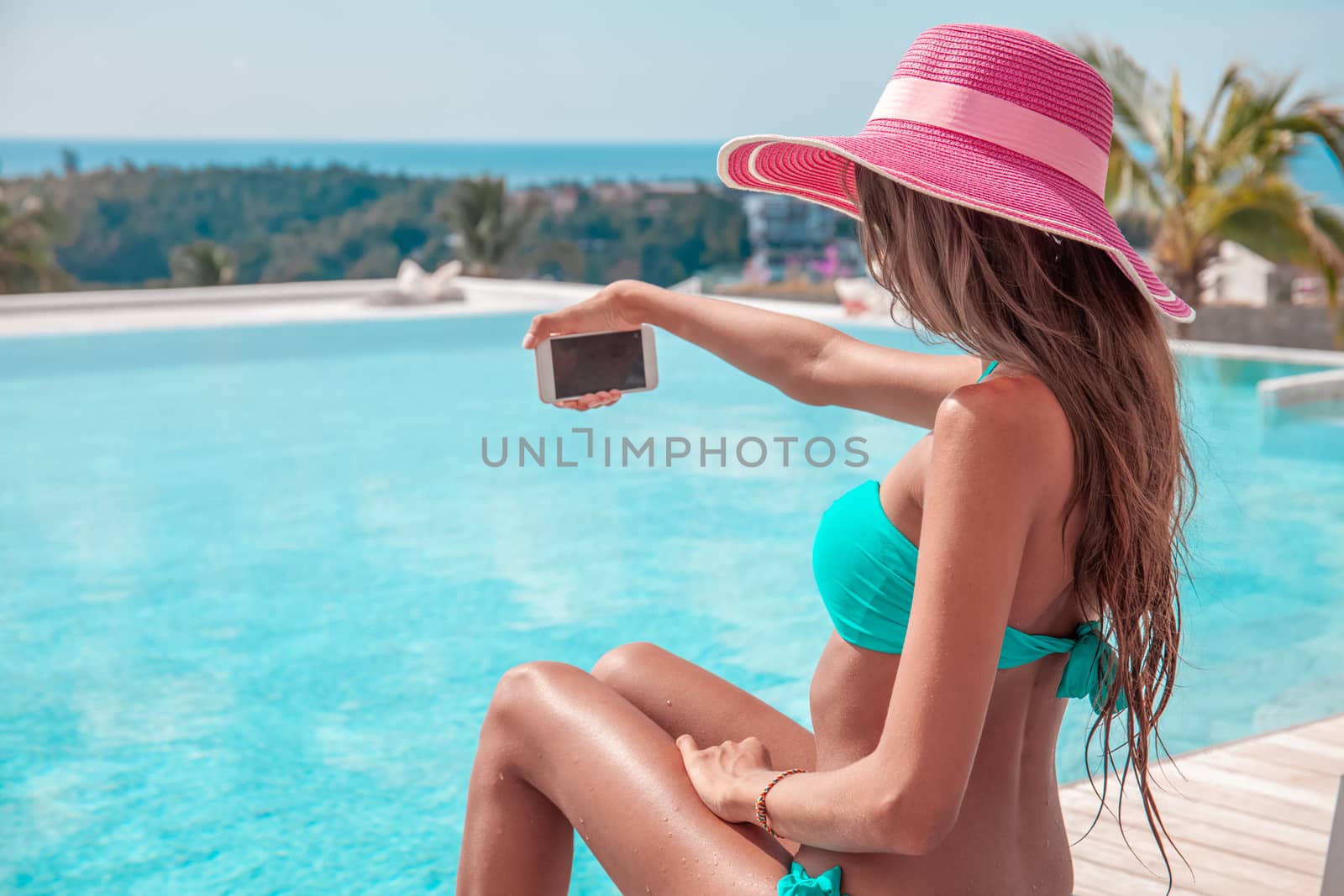 Portrait of a beautiful woman in bikini and sunhat making selfie photo on smartphone outdoors sitting at tourist resort by the pool