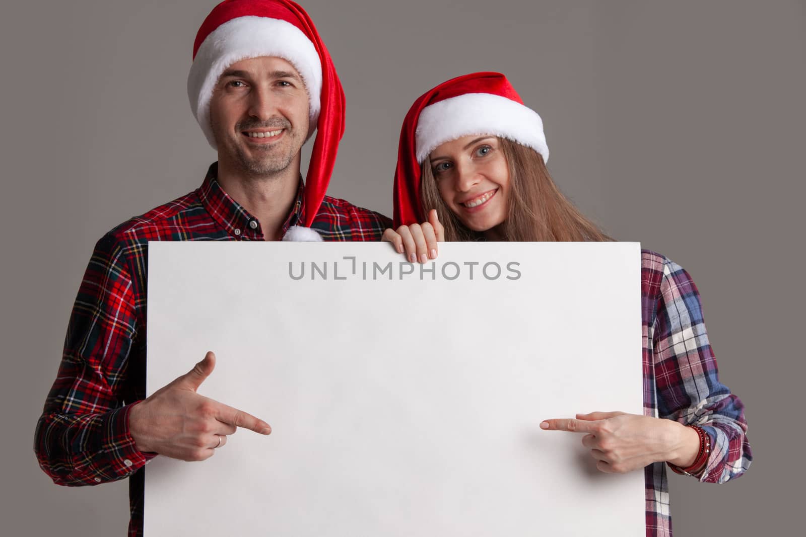 Happy couple in Santa hats holding blank banner and pointing at copy space