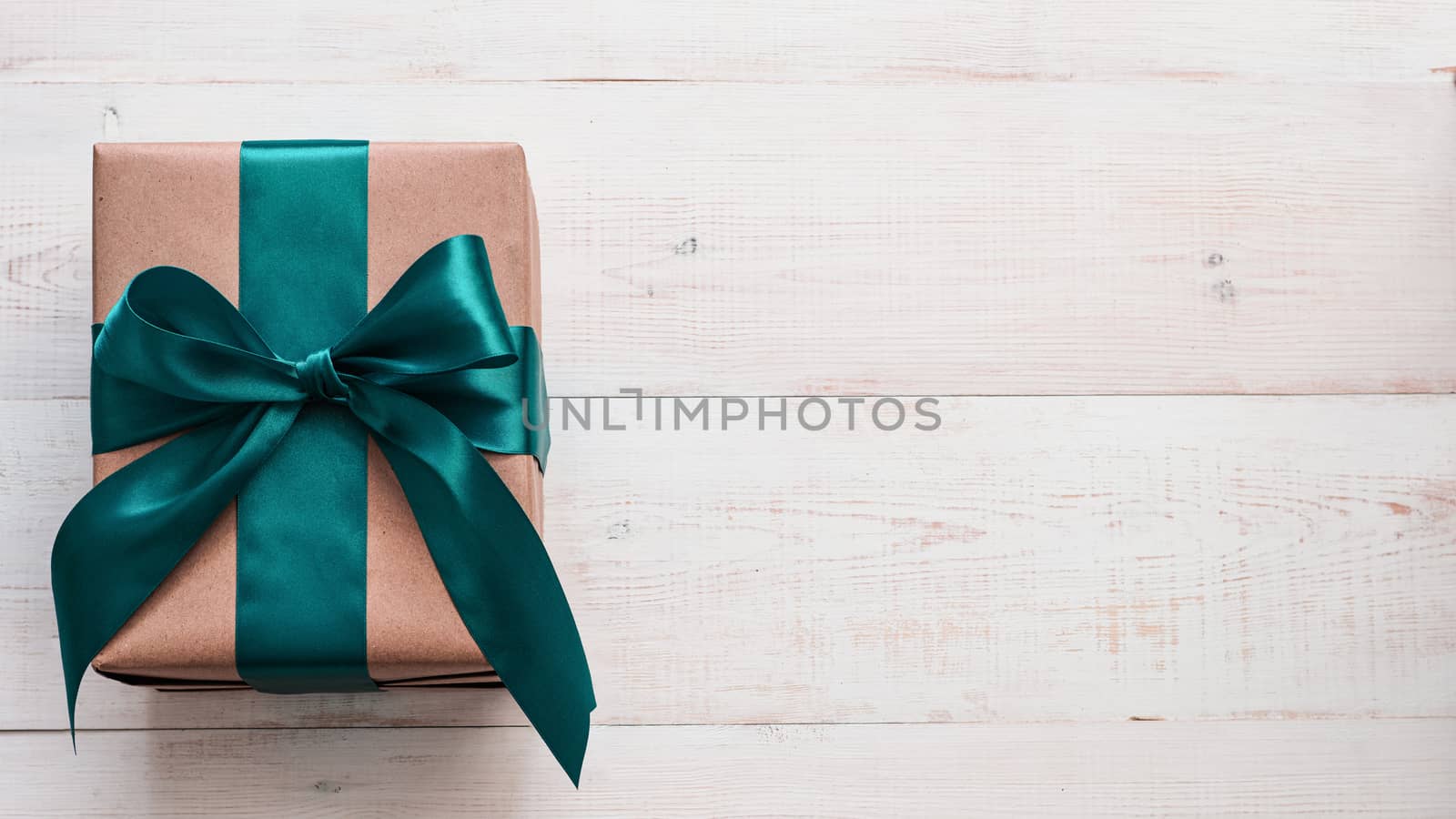 Gift box in craft wrapping paper and green satin ribbon on white wooden table, copy space right. Beautiful Christmas, New Year or Birthday present, flat lay or top view. Banner