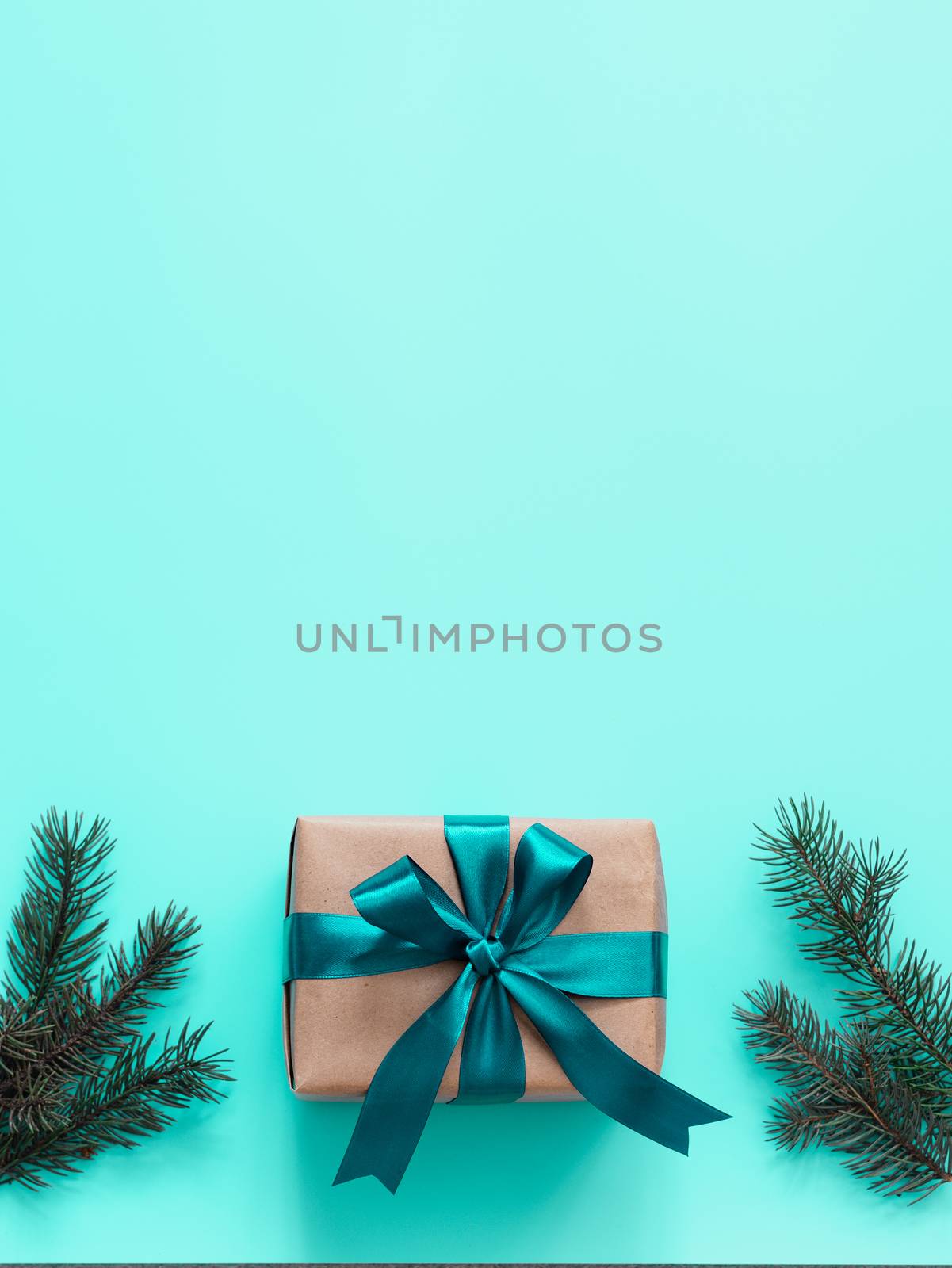 Gift box in craft wrapping paper and green satin ribbon on turquoise blue background, copy space top. Beautiful Christmas or New Year present and fir branches, flat lay or top view. Vertical