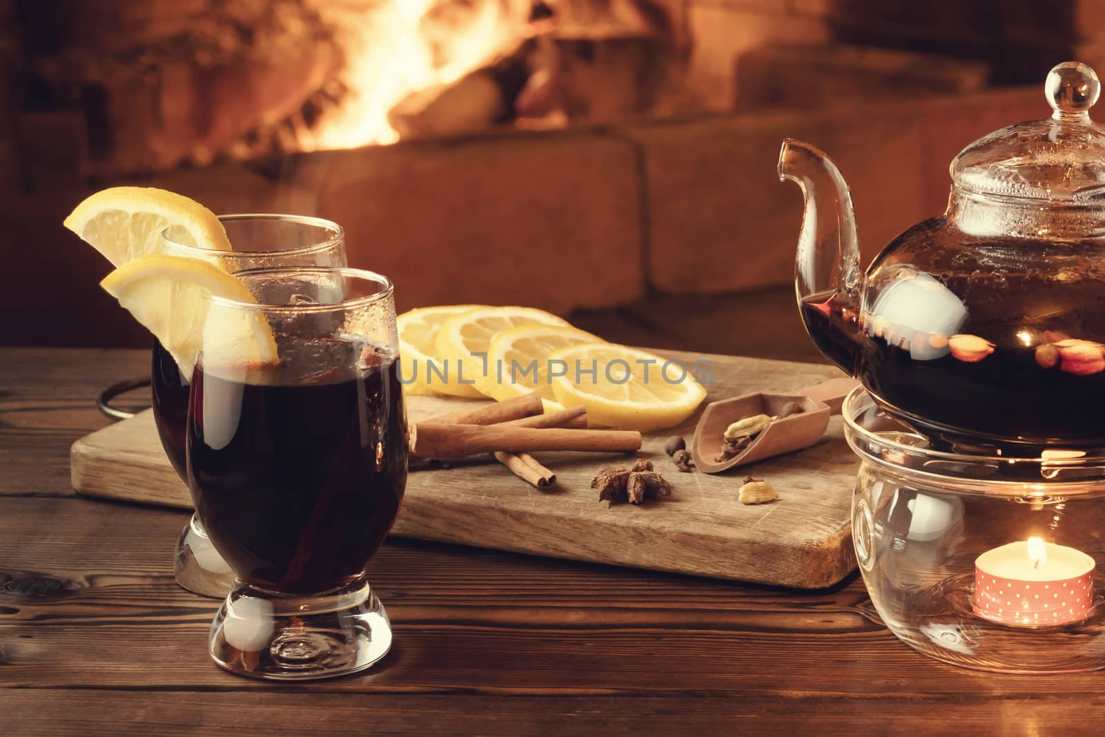 Two glasses of mulled wine and teapot on a wooden table in front of a burning fireplace.