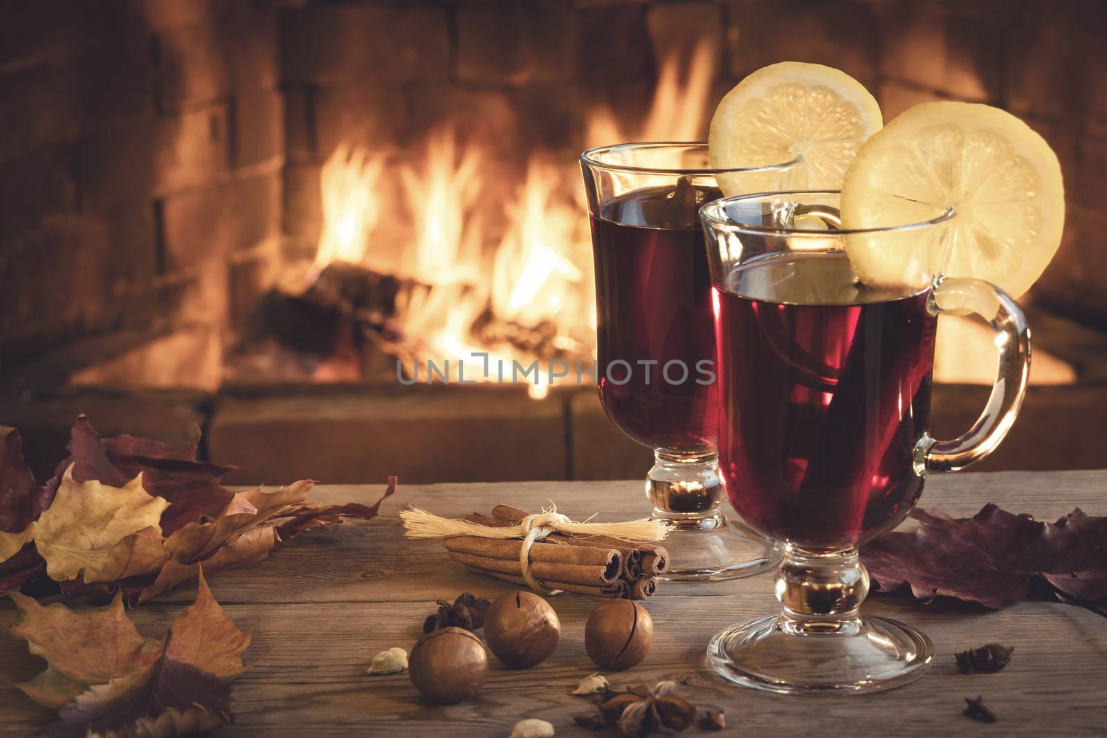 Two glasses of mulled wine on a wooden table in front of a burning fireplace.