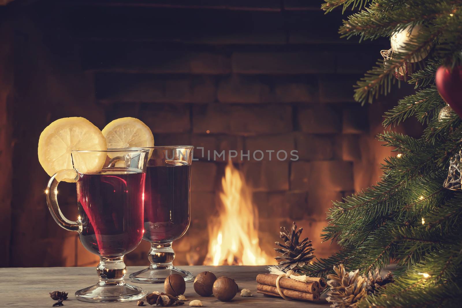 Two glasses of mulled wine on a wooden table near a Christmas tree in front of a burning fireplace.