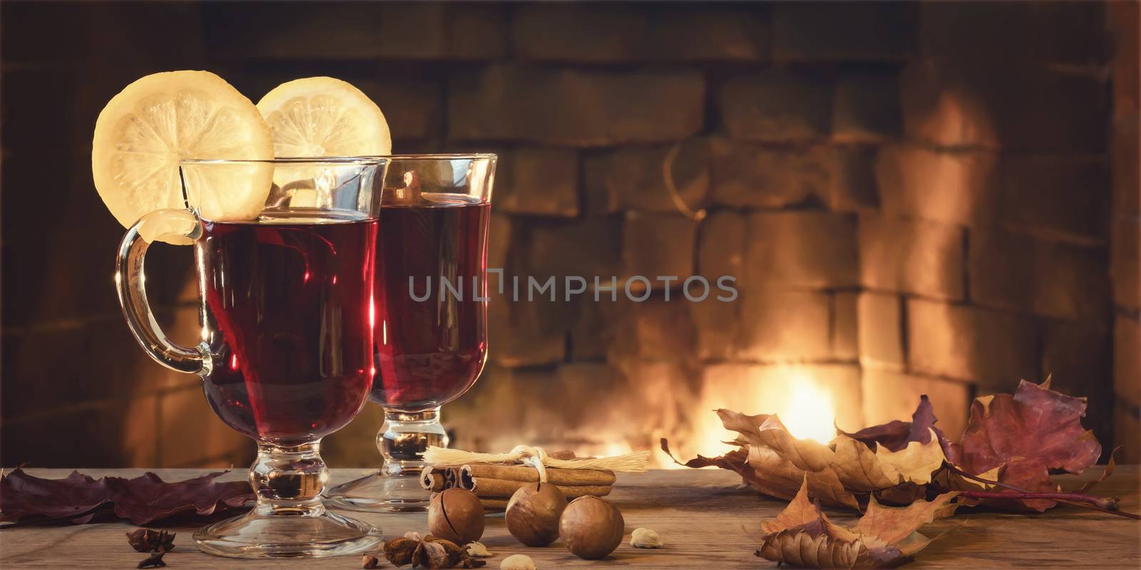 Two glasses of mulled wine on a wooden table in front of a burning fireplace, horizontal banner, copy space.
