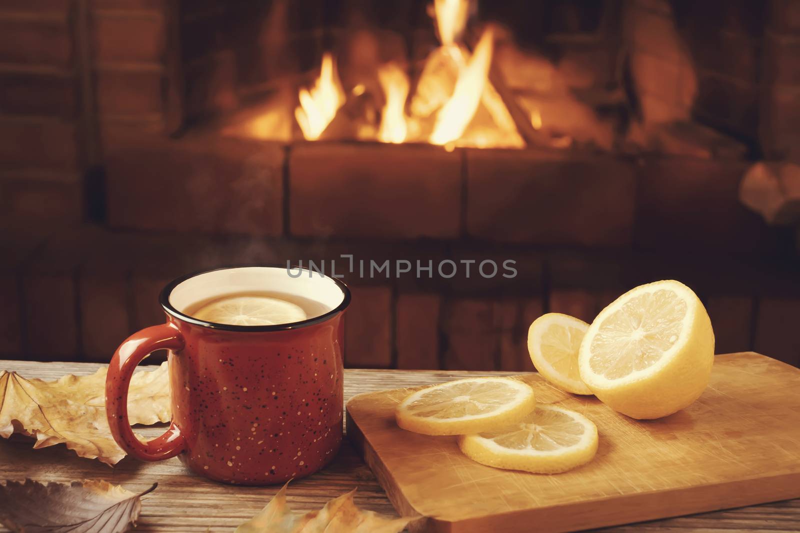 Red mug with hot tea with lemon in front of a burning fireplace, comfort and warmth of the hearth concept.