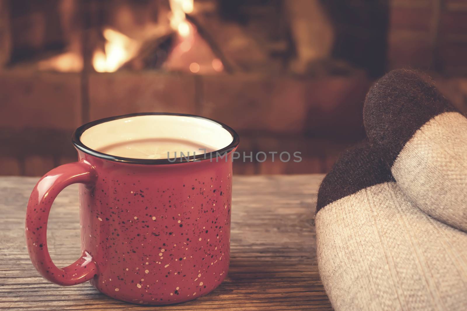 Feet in woolen socks, a red mug with hot tea opposite the burning fireplace, comfort, and warmth of the hearth concept by galsand