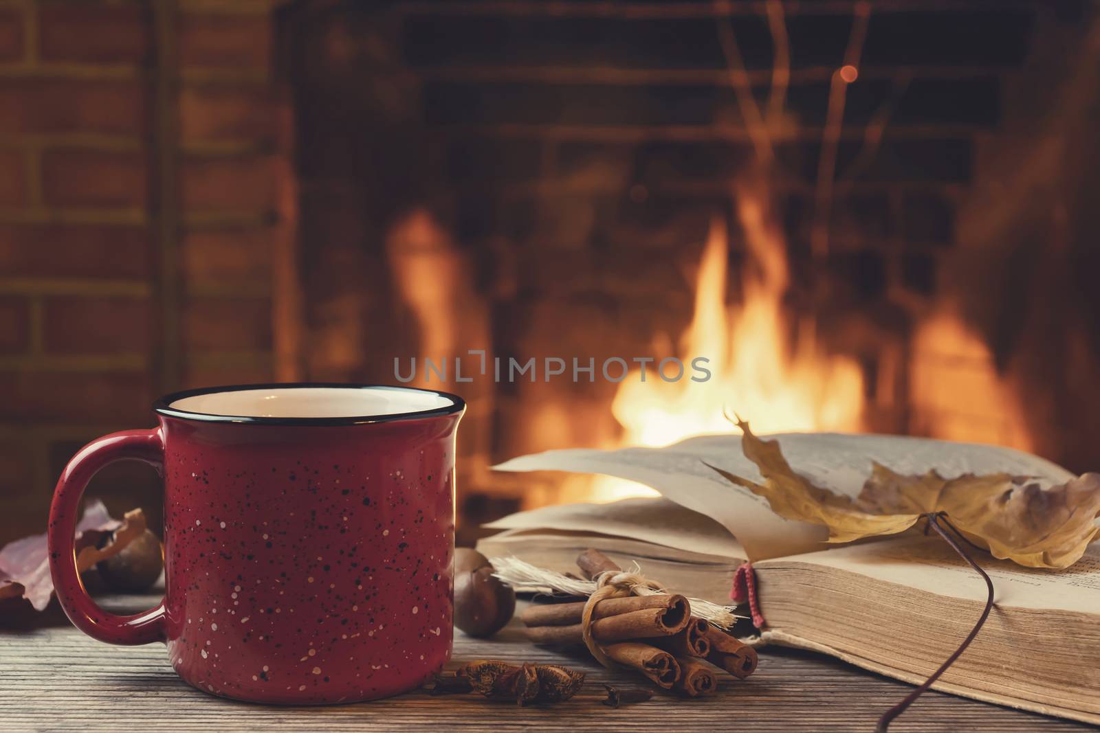 Red mug with hot tea and an open book in front of a burning fireplace, comfort, relaxation and warmth of the hearth concept.