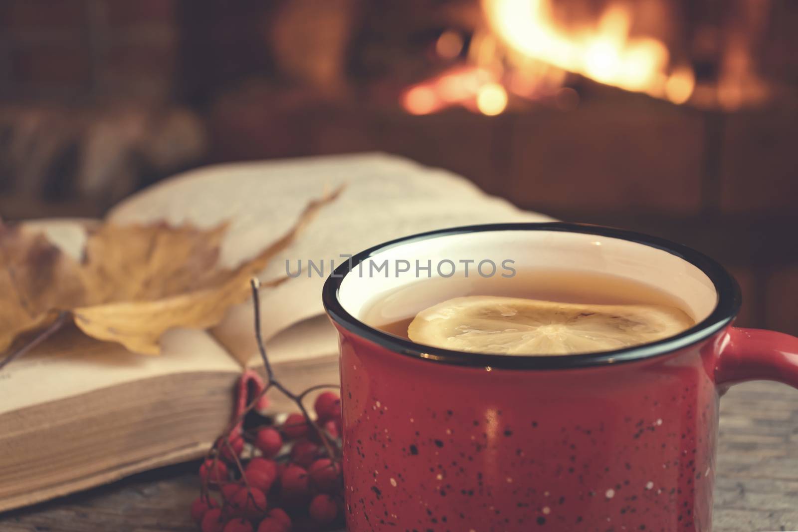 Red mug with hot tea and an open book in front of a burning fireplace, comfort, relaxation and warmth of the hearth concept.