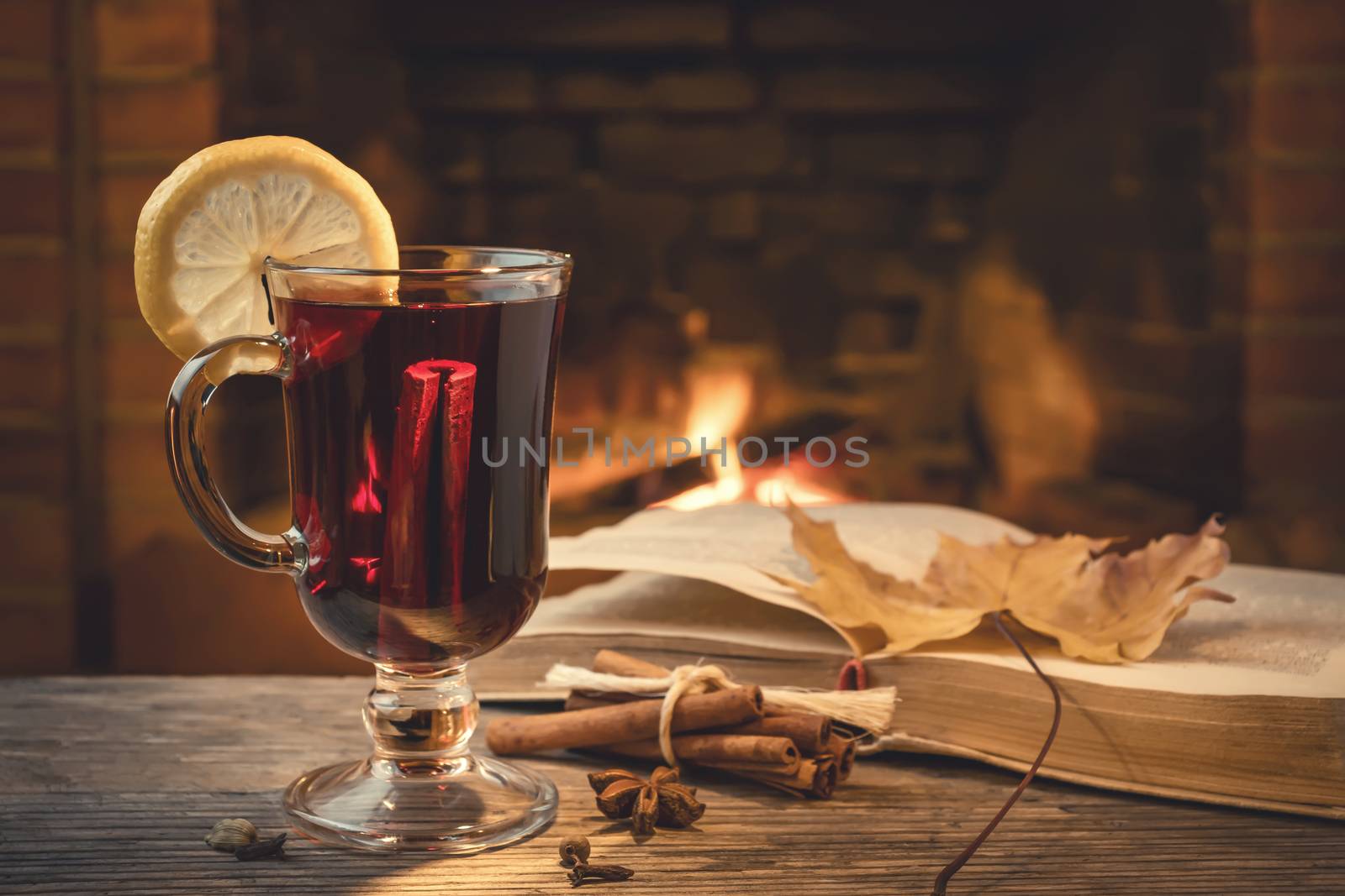 Glass of mulled wine, spices, a book on a table in a cozy room with a burning fireplace by galsand