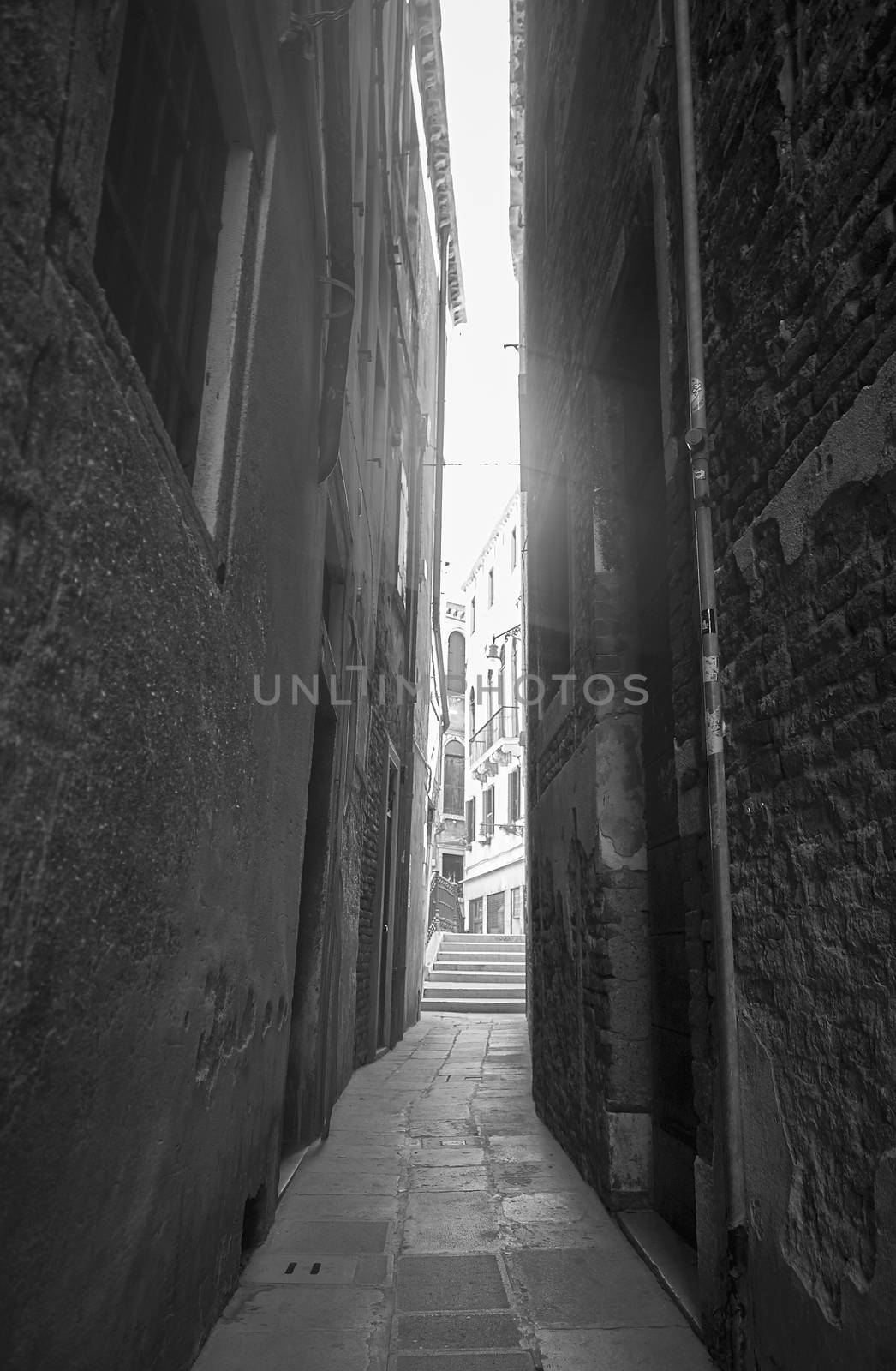 A very narrow empty alley with brick walls and a strong light in front of the sky. Black and white image.