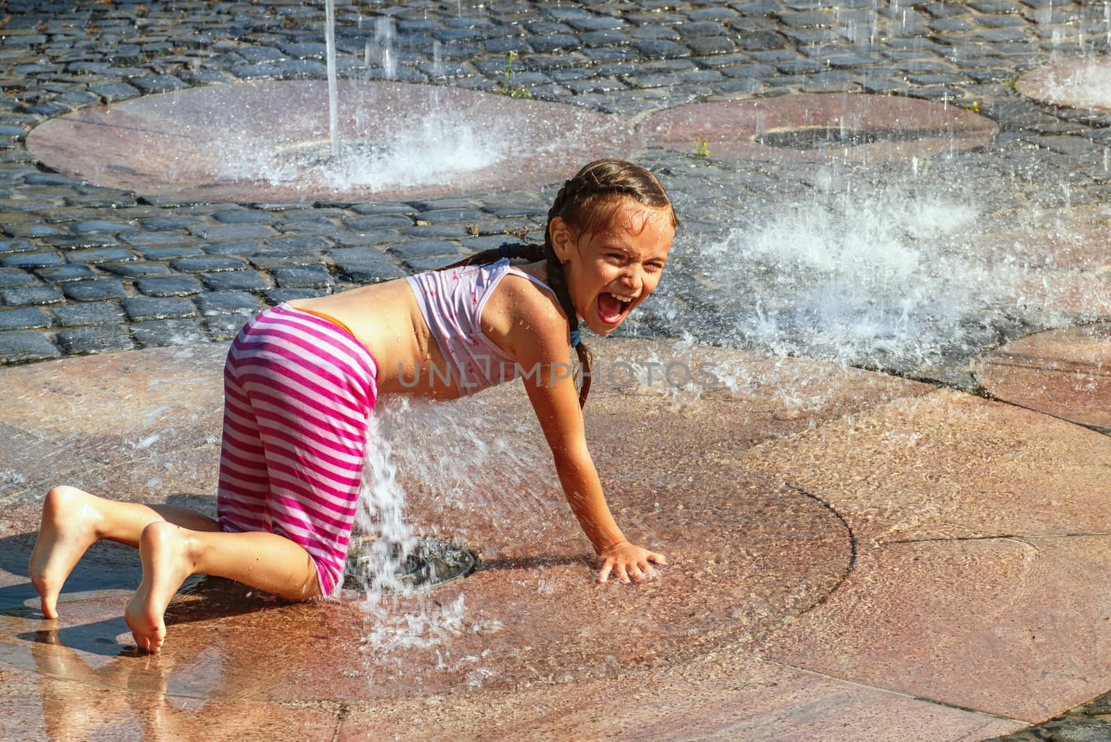 Girl on a sunny summer day are poured water from a fountain.Girl happily in shallow clean water on of city fountain on warm bright summer day.