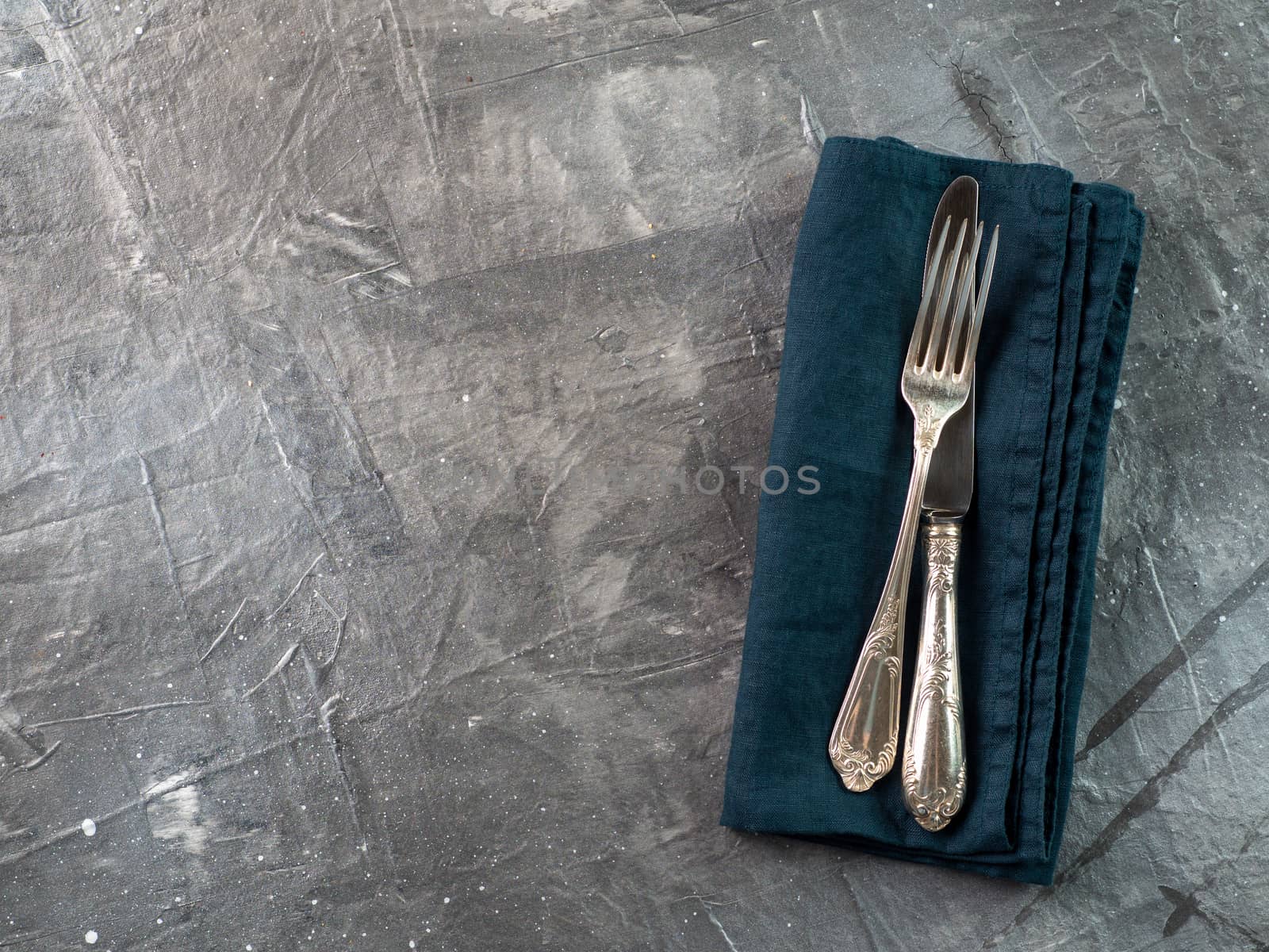 Silver fork and knife on dark green emerald linen napkin over gray textured background table with copy space. Cutlery with copy space, top view or flat lay