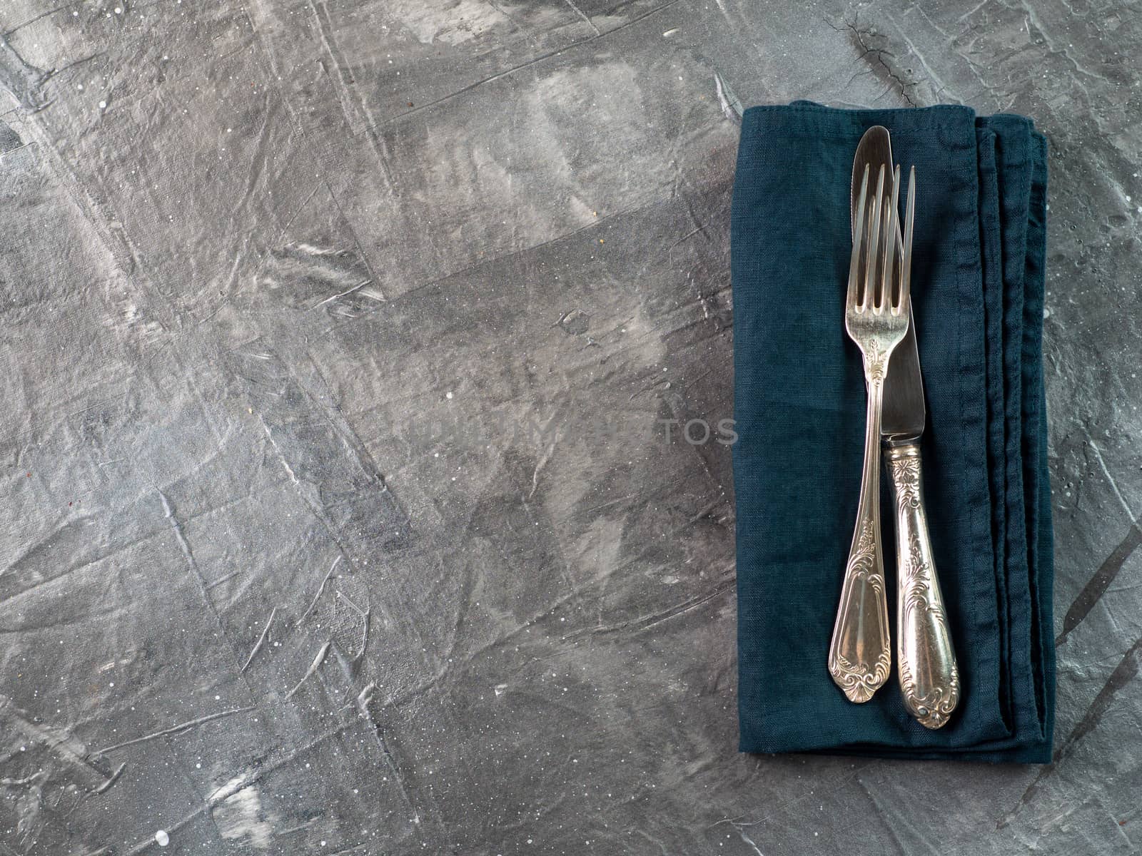 Silver fork and knife on dark green emerald linen napkin over gray textured background table with copy space. Cutlery with copy space, top view or flat lay