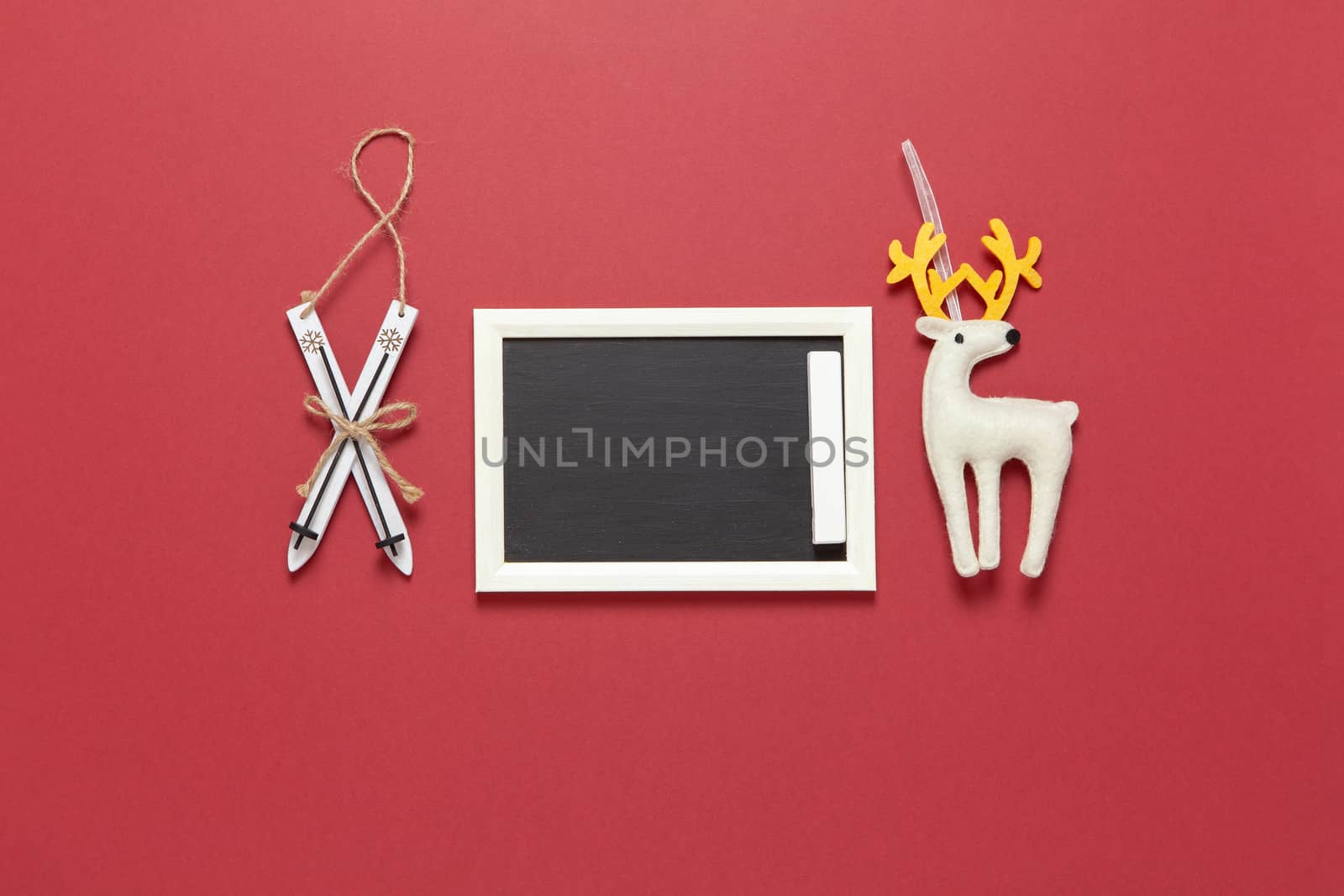 Christmas toys wooden skis, white felt toy deer and small clean chalk board with copy space on dark red background. Festive, New Year concept. Horizontal, flat lay. Minimal style. Top view.