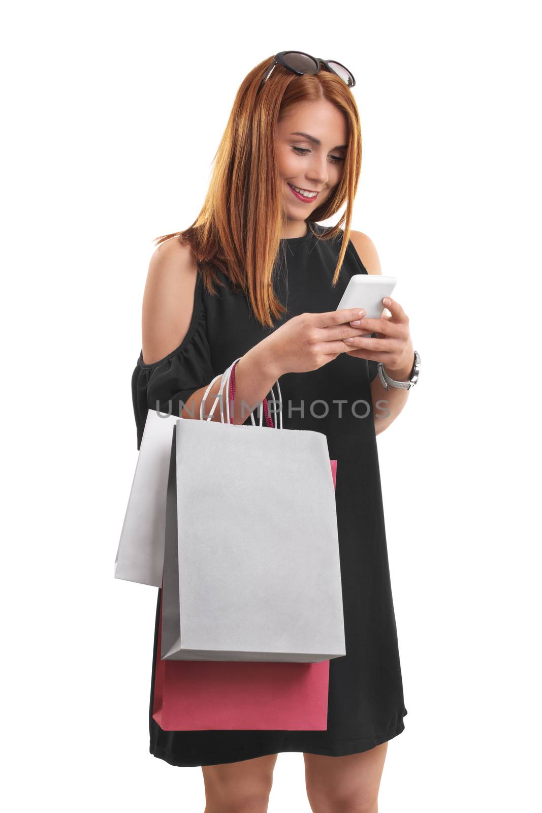 Portrait of a beautiful young girl in a stylish dress looking and typing at her phone, holing shopping bags, isolated on white background.