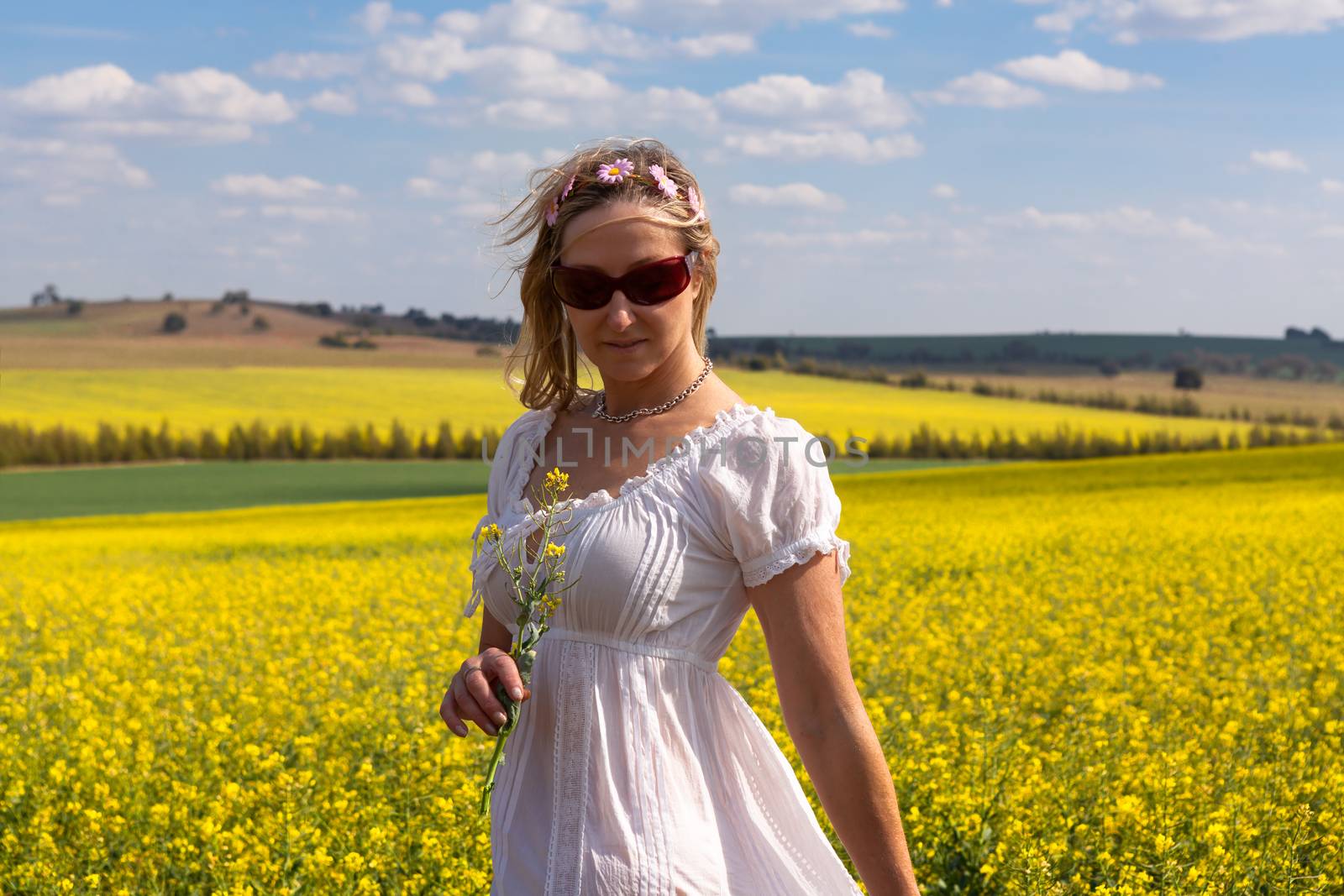 Female in white cotton dress stands by a field of golden canola and rolling agriculural farmland and hills in springtime