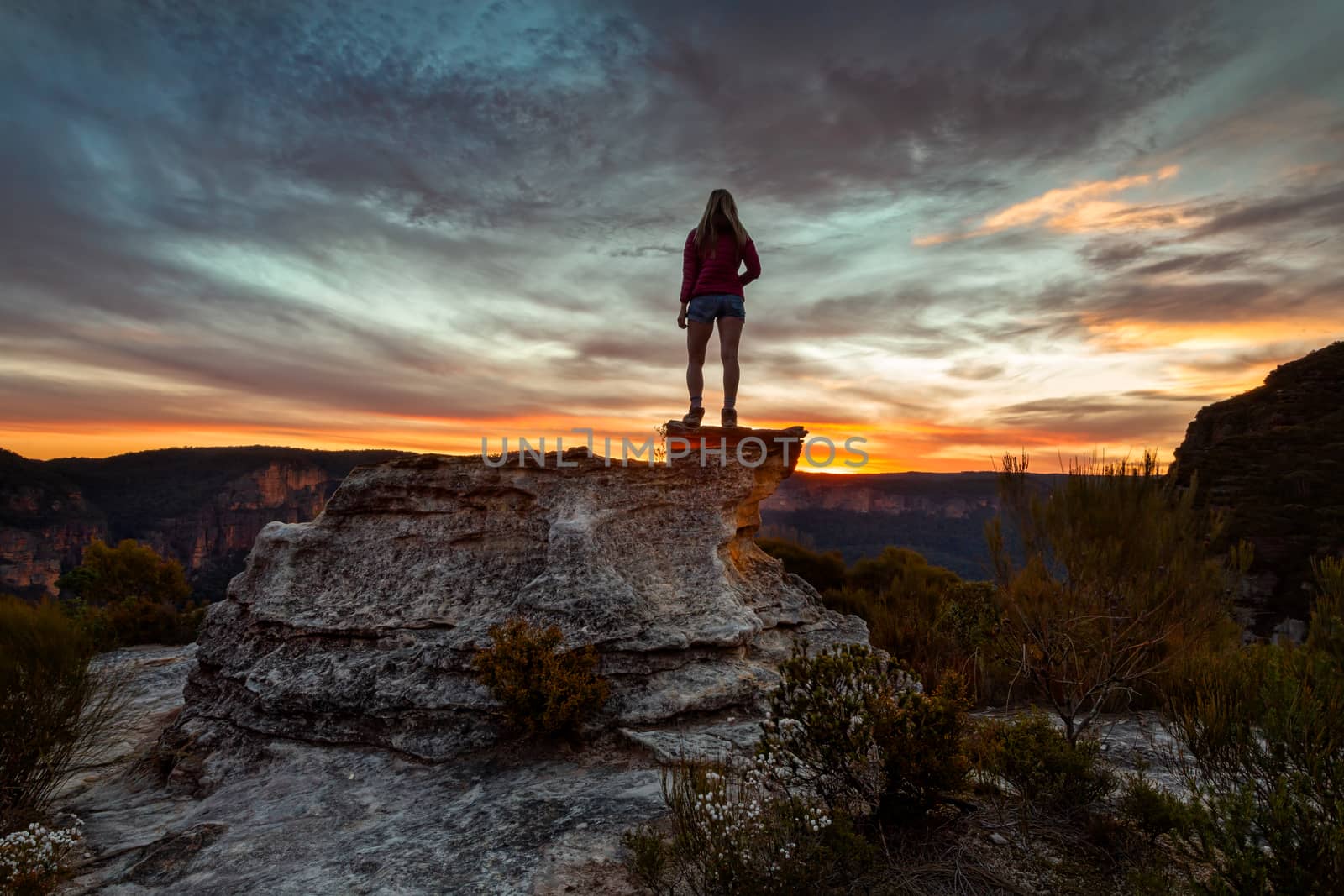 Female hiker stands on mountain peak rocky outcrop along the cliifs of Blue Mountains, taking in spectacular views as the sun sets.