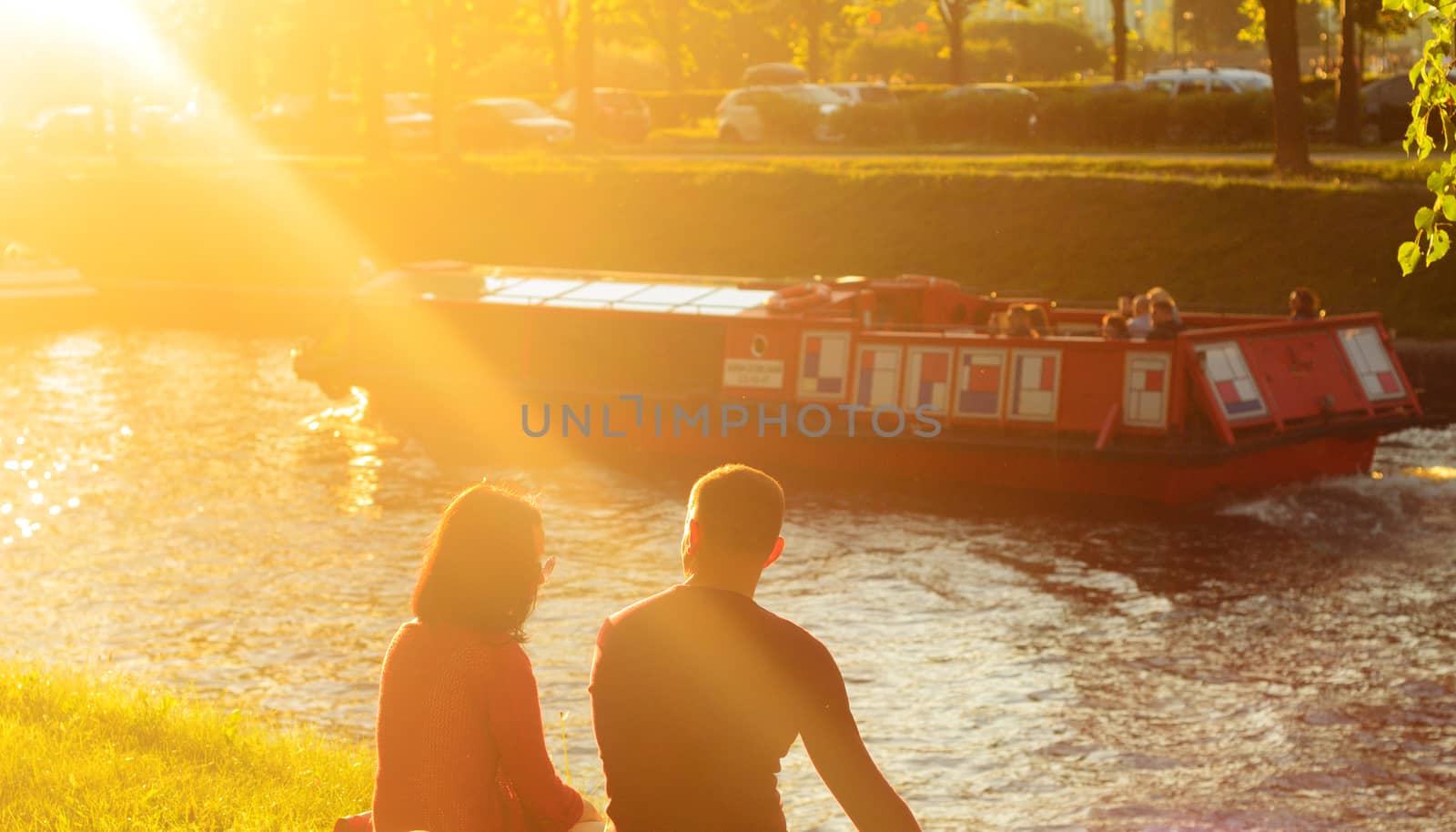 At sunset a lovely couple sits on the bank of a river canal in a city park and talks each other.The canal floats pleasure boat.Good Summer weekend. by andre_dechapelle