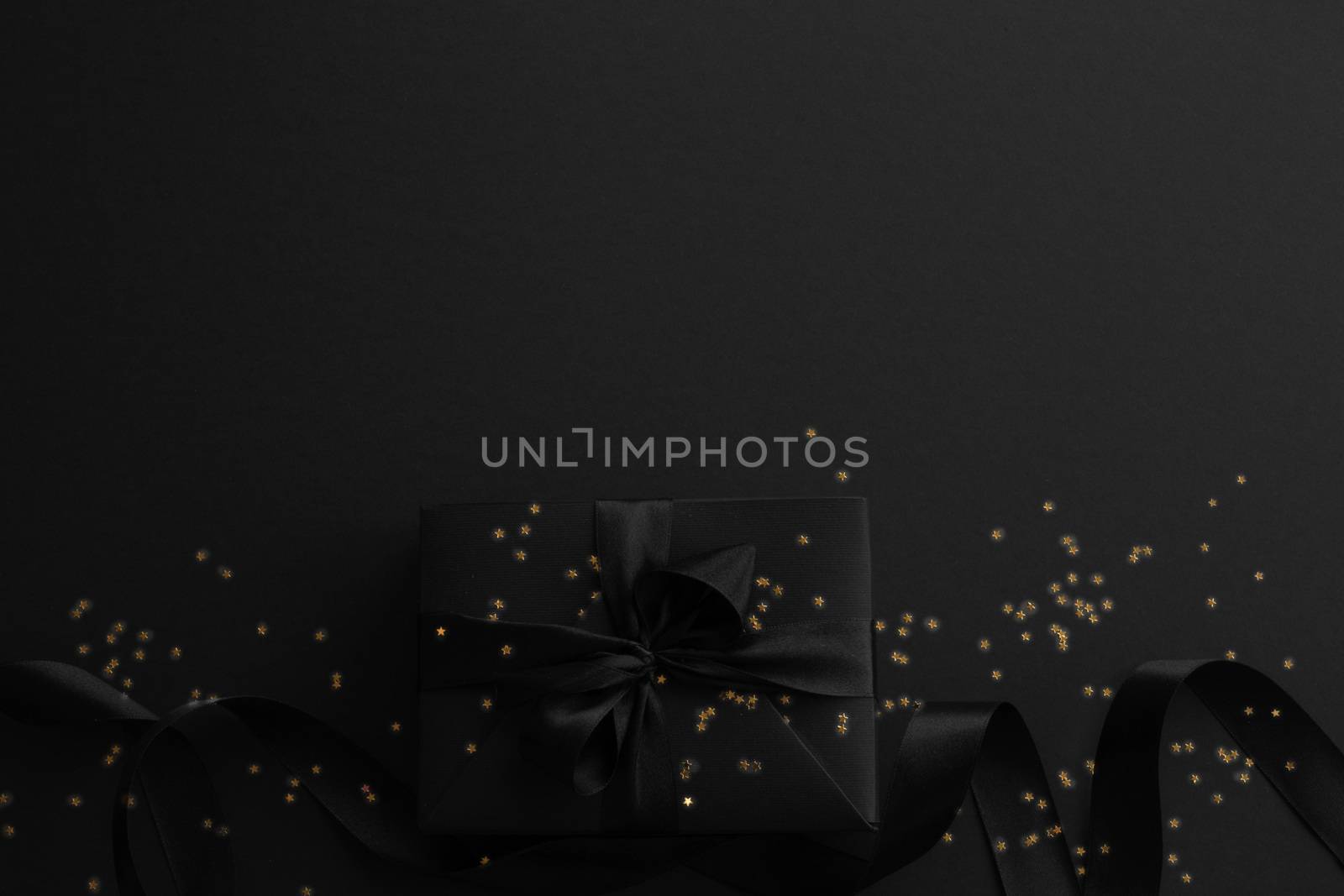 Black friday gift, paper box with silk ribbon bow and golden glitter stars on black paper background with copy space for text, flat lay top view template