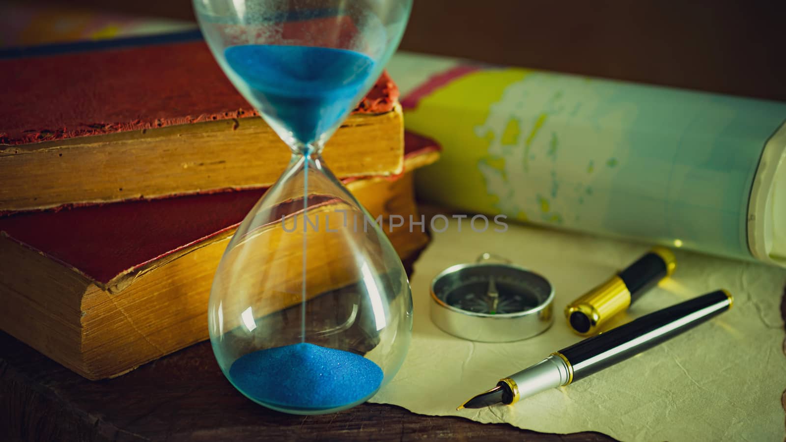 Sand clock with old books and pen with paper map on the wooden table. Concept of travel planning.