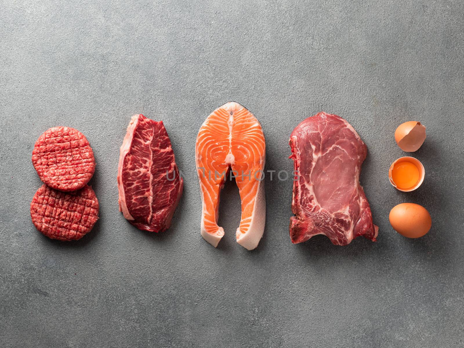 Carnivore or keto diet concept. Raw ingredients for zero carb or low carb diet - burger patties, ribeye, salmon steak, pork, egg on gray stone background.Top view or flat lay.Copy space top and bottom