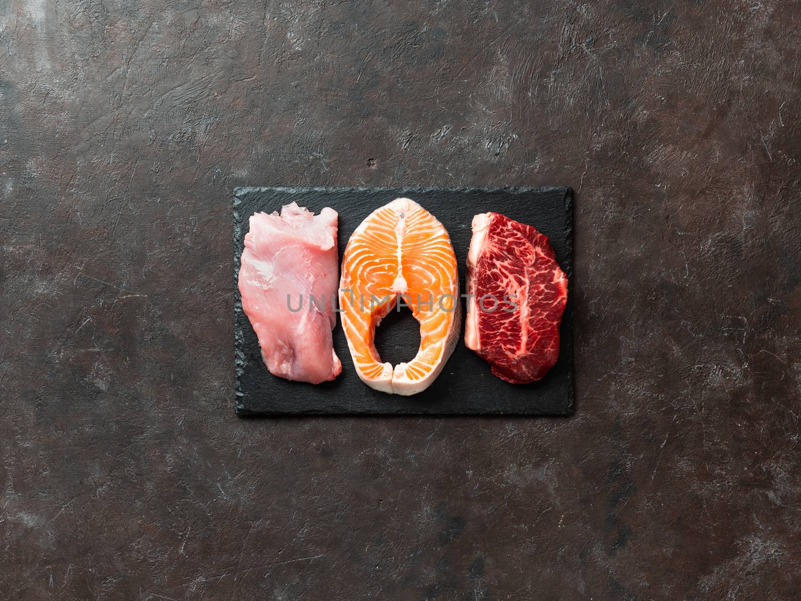 Raw turkey breast fillet, salmon steak and beef ribeye steak on black slate board over dark background. Top view or flat lay. Protein food, keto, carnivore, paleo diet concept. Copy space.