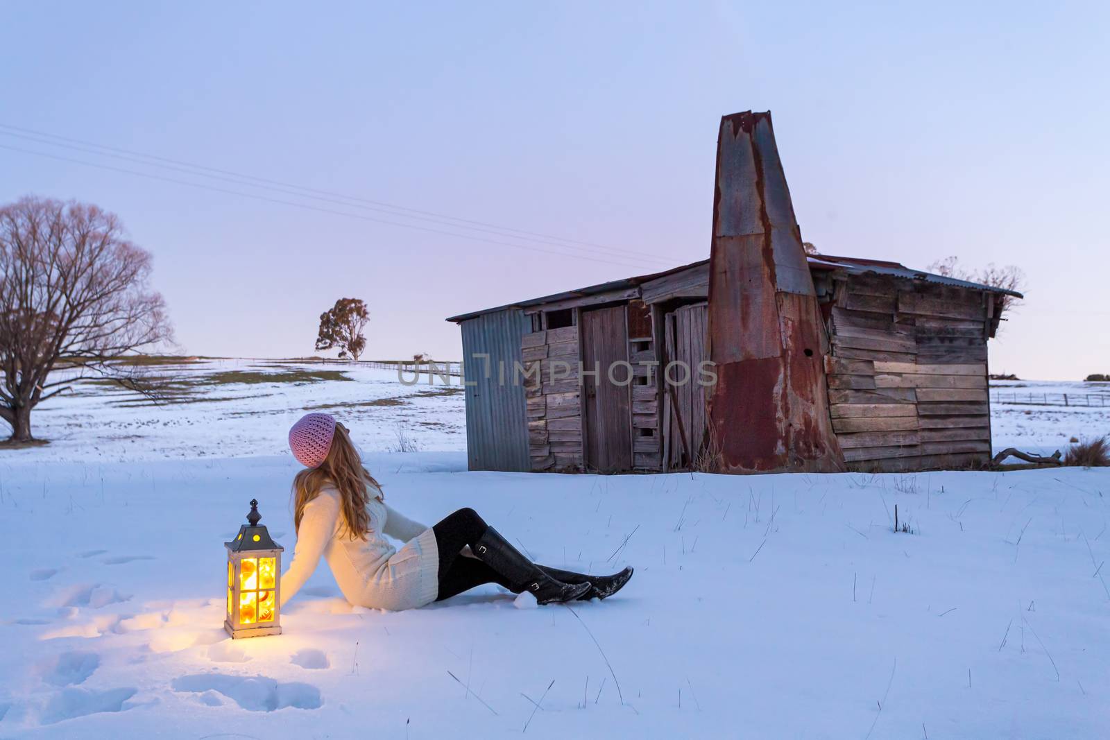 Woman with lantern sitting in a snow covered field.  The day is drawing to a close and the cool dusk colours envelop the landscape.  A rustic stable shed  of mismatched timber and metal nearby