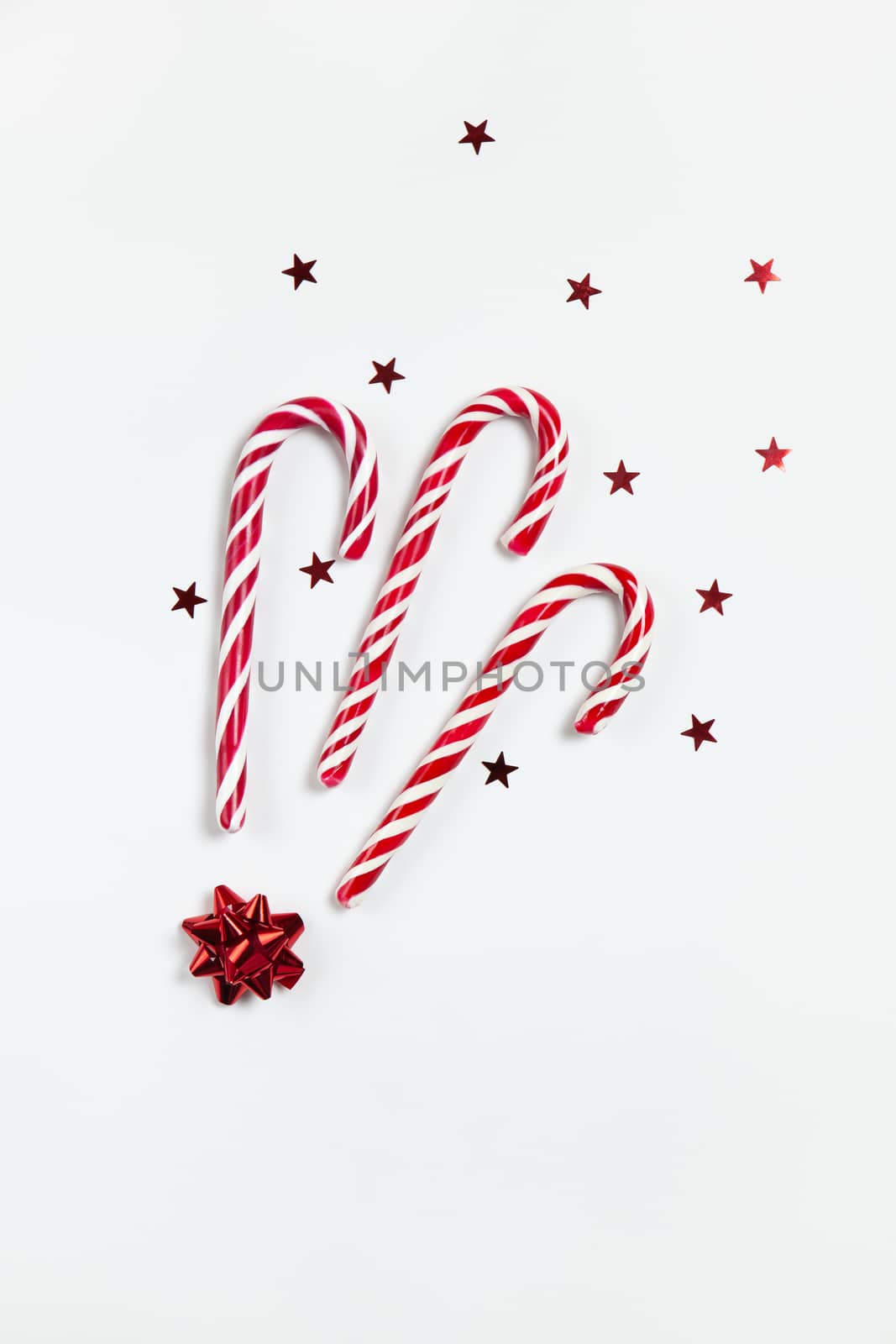 Christmas composition three caramel candy canes, confetti stars and red bow on white background. Festive minimal style flat lay. For greeting card, invitation, social media. Vertical.