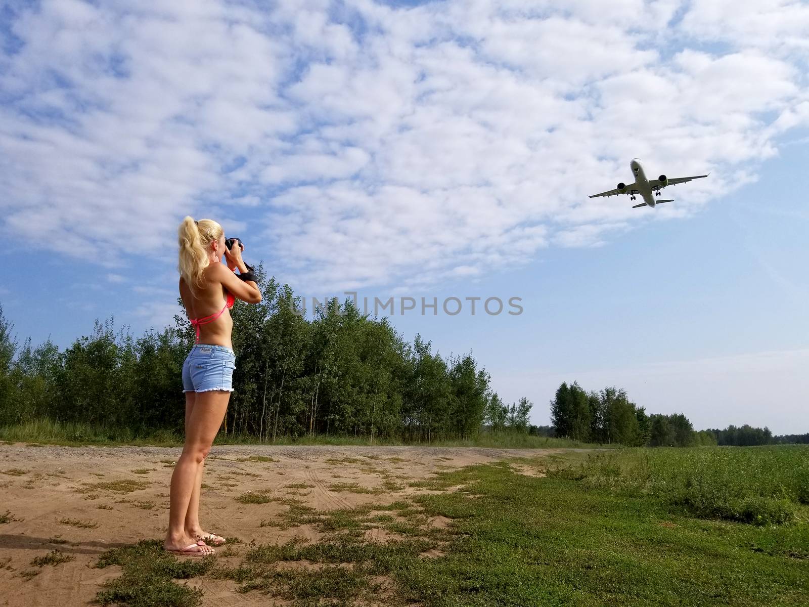 Blonde girl with long hair takes pictures of a plane flying over her on a cloudy sky background.