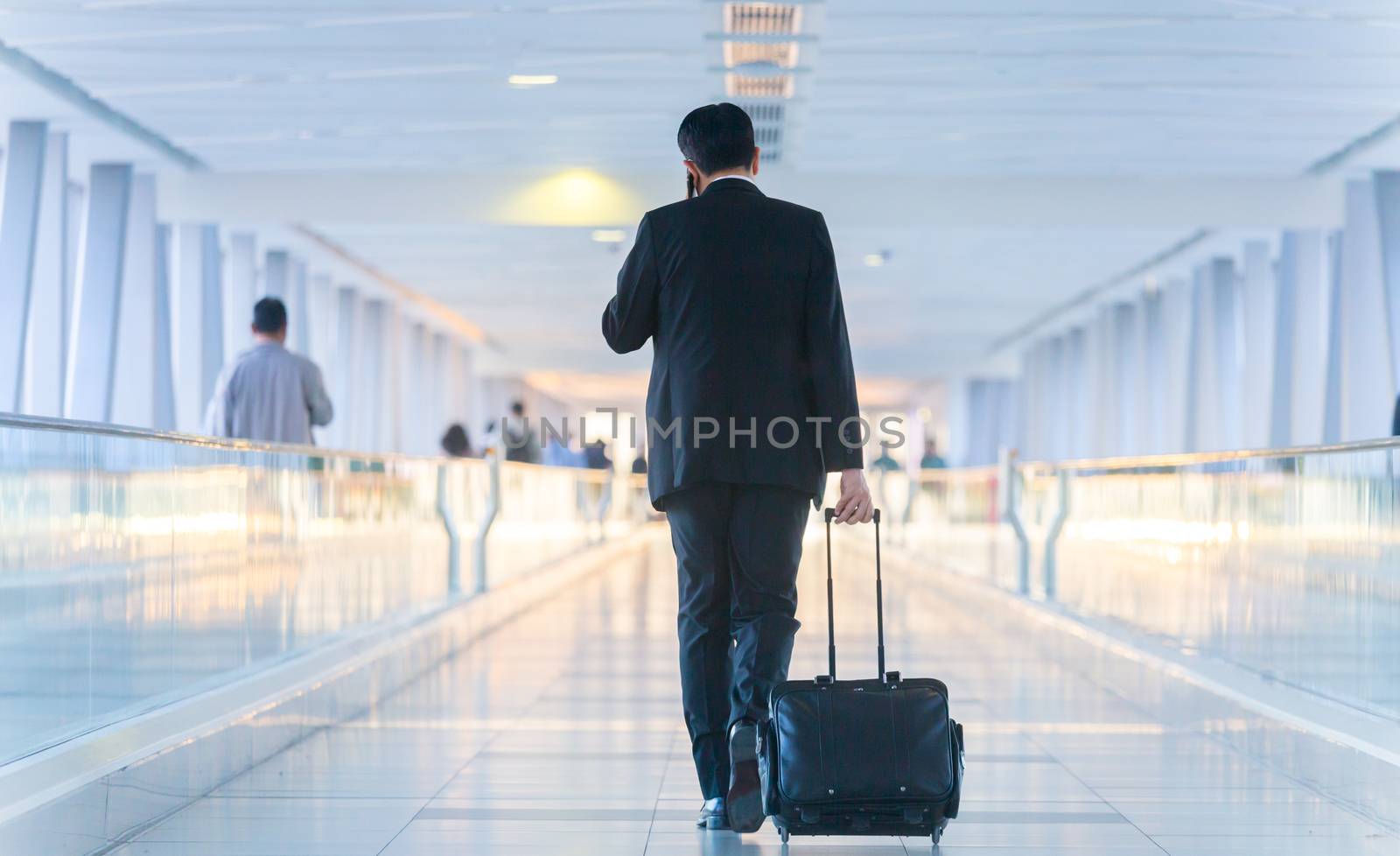 Businessman walking and wheeling a trolley suitcase at the lobby, talking on a mobile phone. Business travel concept. by kasto