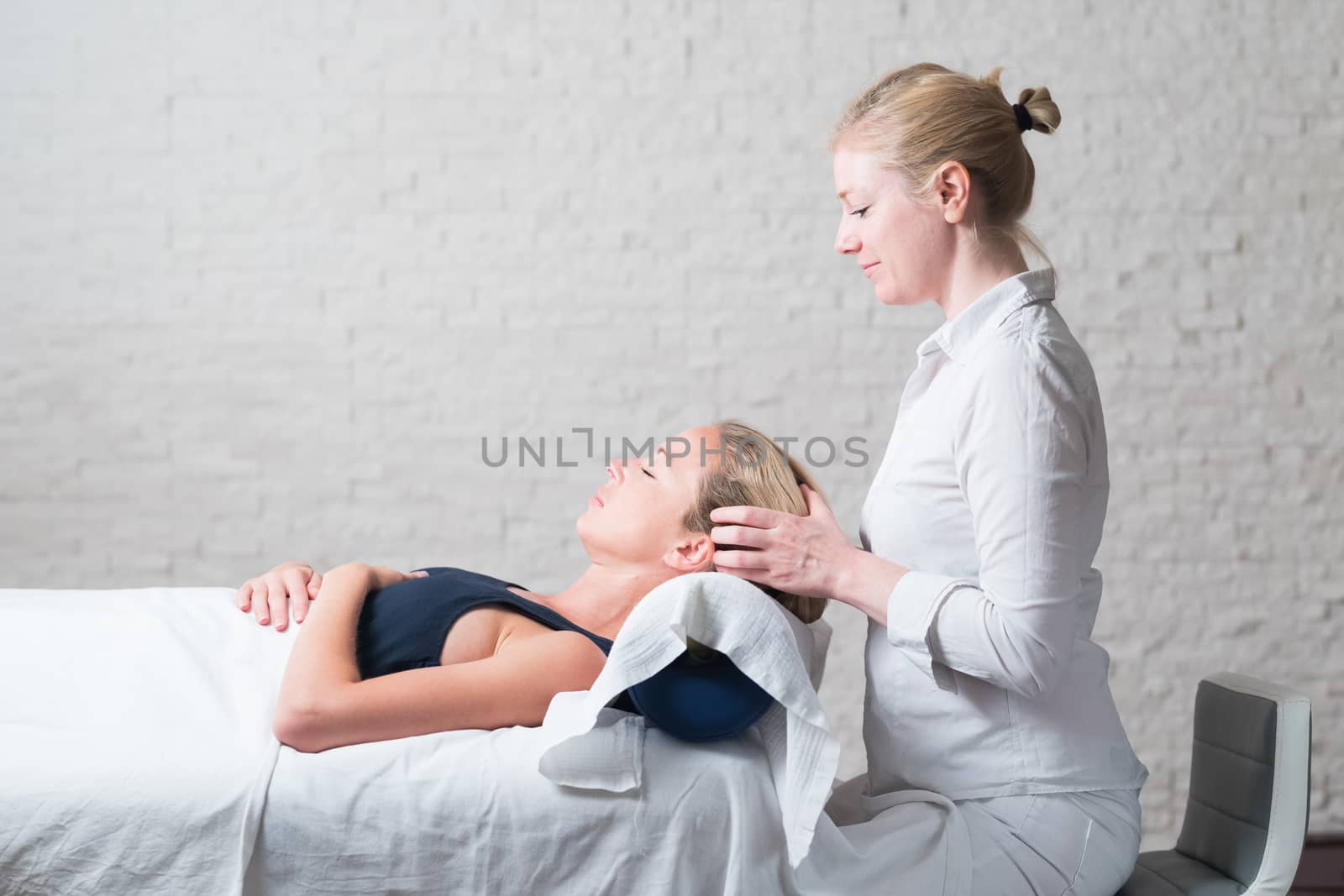 Professional female masseur giving relaxing massage treatment to young female client. Hands of masseuse on forehead of young lady during procedure of spa facial massage.