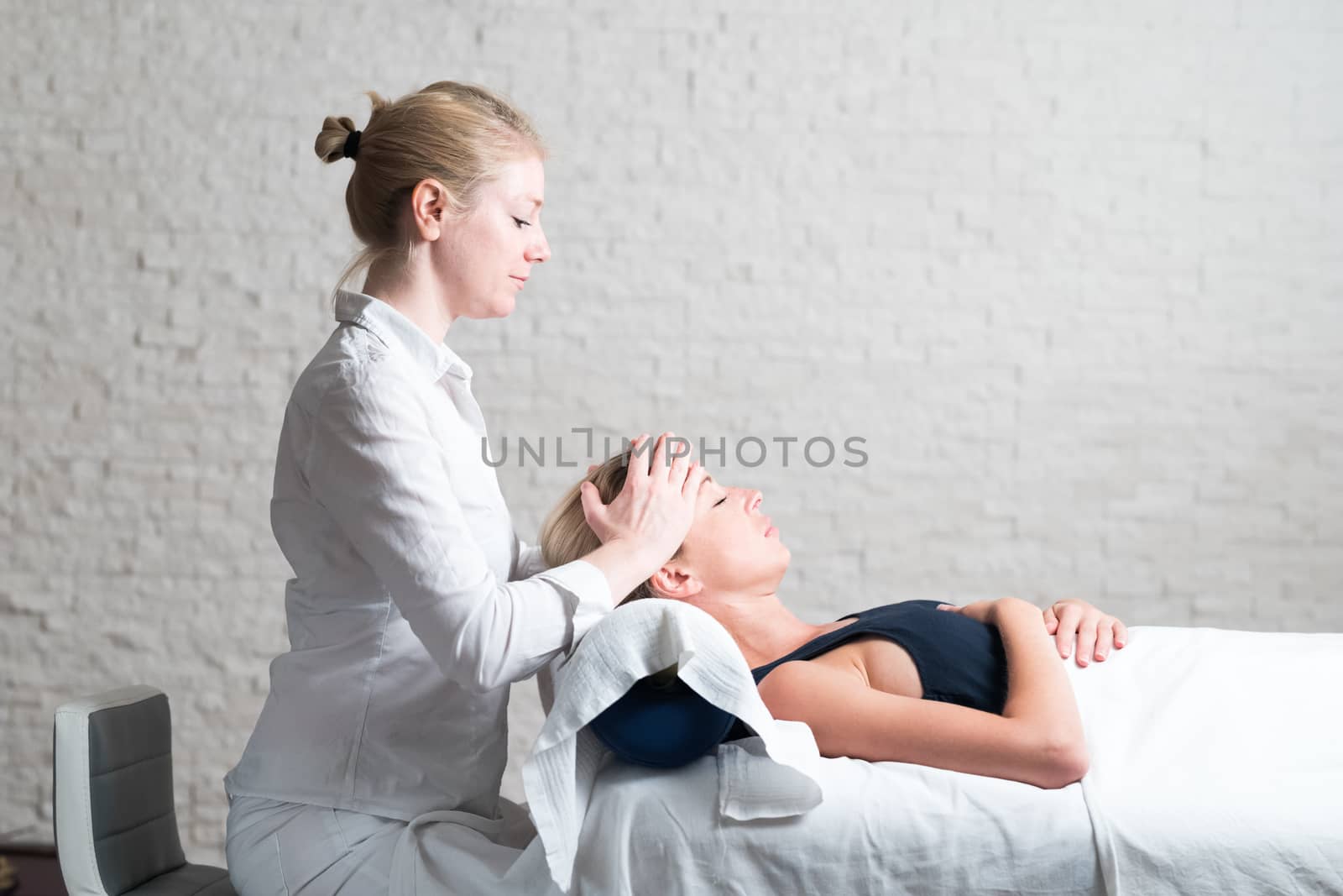 Professional female masseur giving relaxing massage treatment to young female client. Hands of masseuse on forehead of young lady during procedure of spa facial massage by kasto