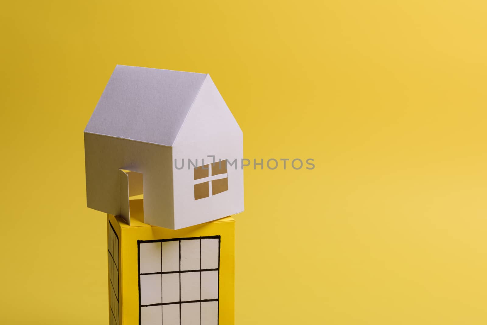 White family paper house over block of flats on yellow background paper. Minimalistic and simple concept, style. Copy space. Vertical orientation.