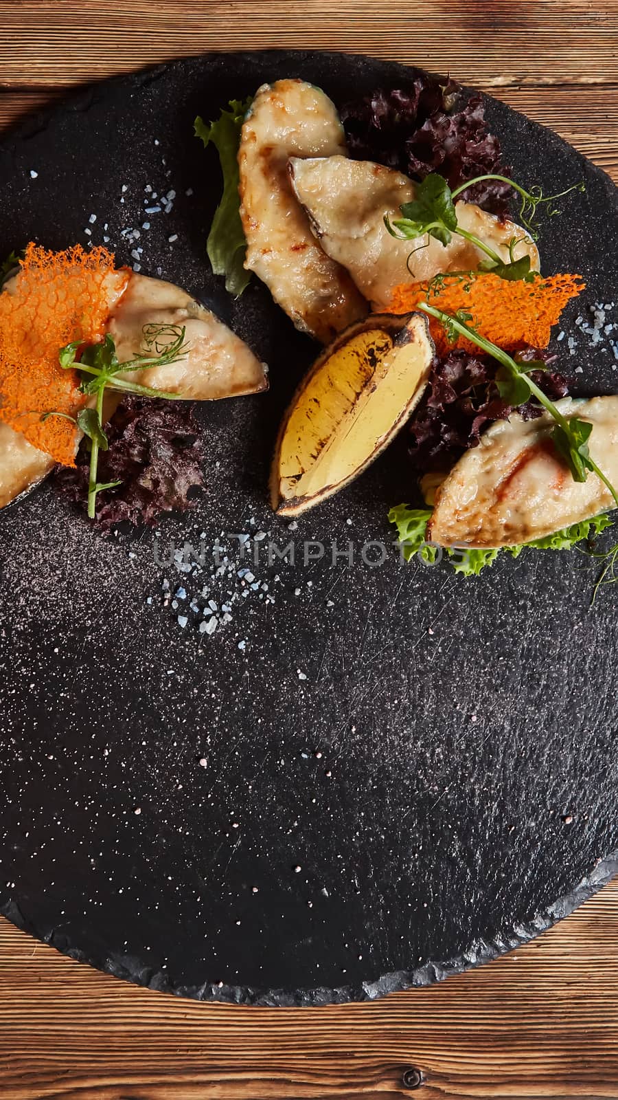 Seafood. Shellfish mussels. Baked mussels with cheese, cilantro and lemon in shells on slate stone board. Plate of mussels, cold white wine, lemon and cilantro on black stone background. Top view