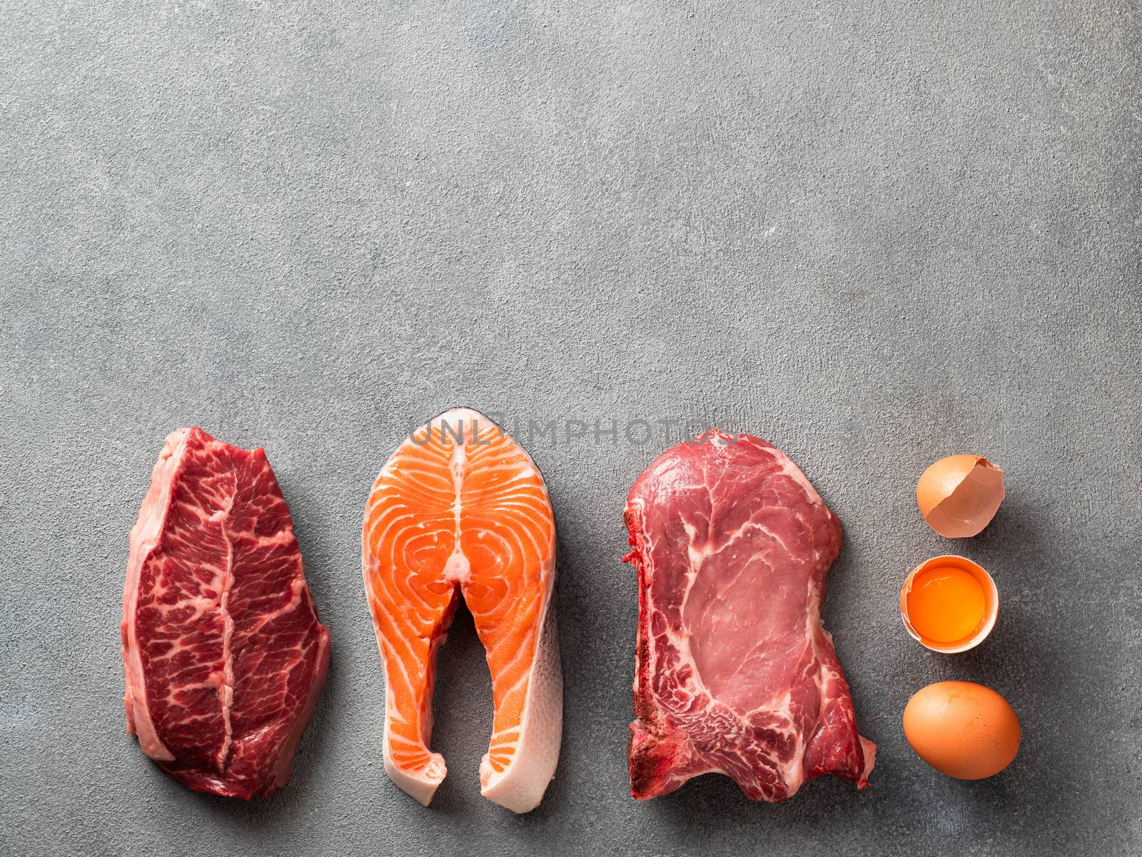 Carnivore or keto diet concept. Raw ingredients for zero carb or low carb diet - rib eye, salmon steak, pork, egg on gray stone background. Top view or flat lay. Copy space top.