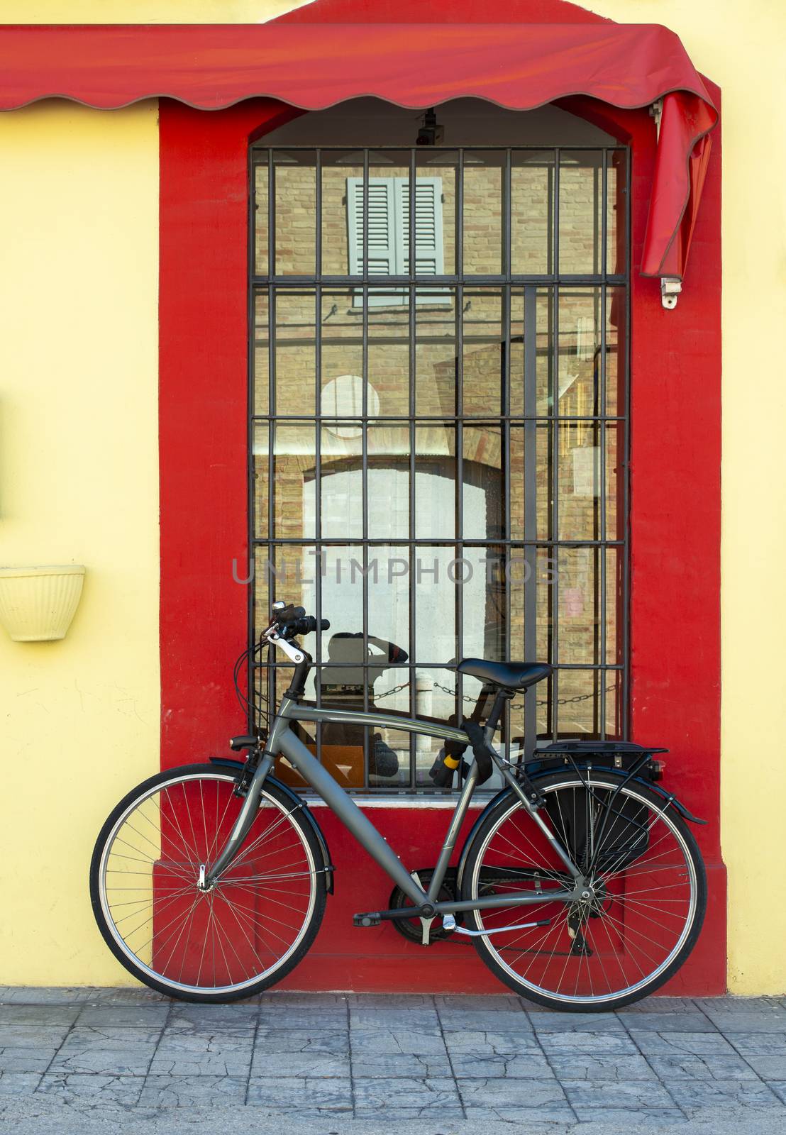 Grey bike in front of yellow facade and red window. Bicycle with trunk. Reflection of old building in the window. Window with grille. 