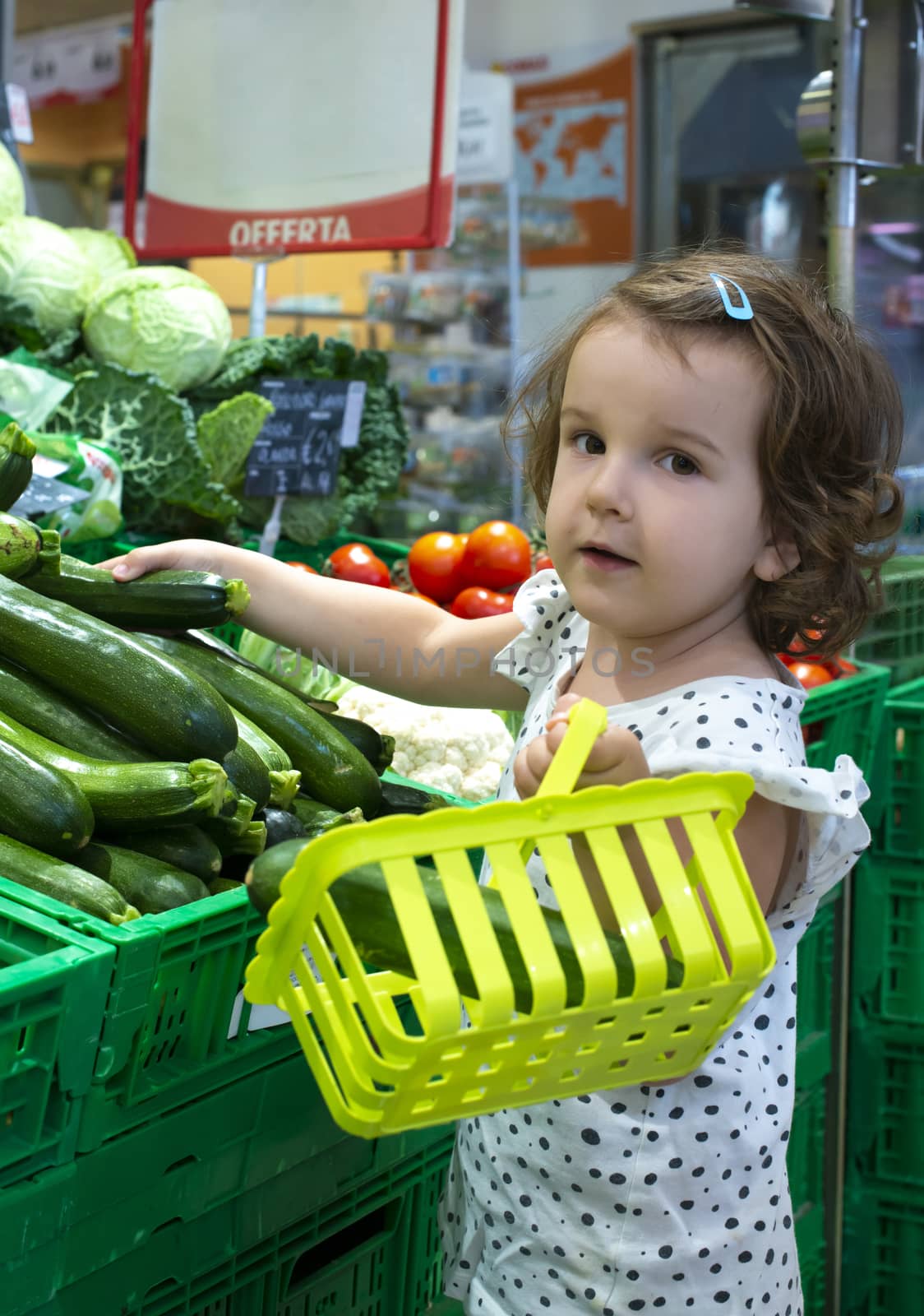 Little girl buying zucchini in supermarket. Looking at the camera. Child hold small basket in supermarket and select vegetables. Concept for healthy eating for children. 