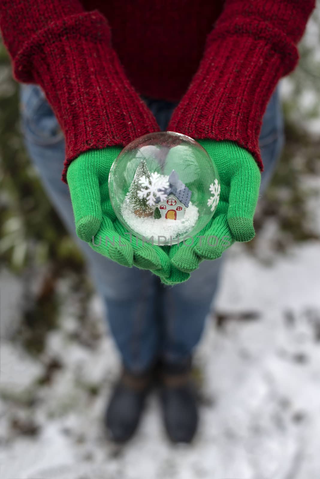 Crystal christmas ball with house and snow inside. Hands with green gloves hold transparent ball.