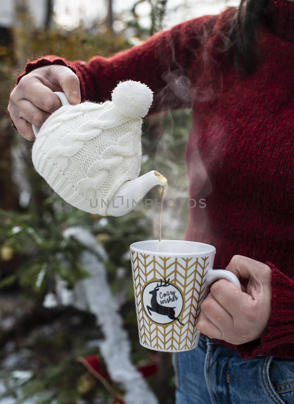 Woman pours tea with a teapot into a teacup. Christmas tree in the foreground. Teacup with Christmas ornaments. Steam comes out of cup of tea. Winter and Christmas concept. Exterior shot.