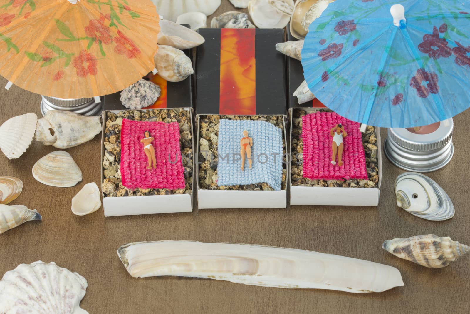 small miniature dolls are sunbathing in matchboxes under the parasol with shell around