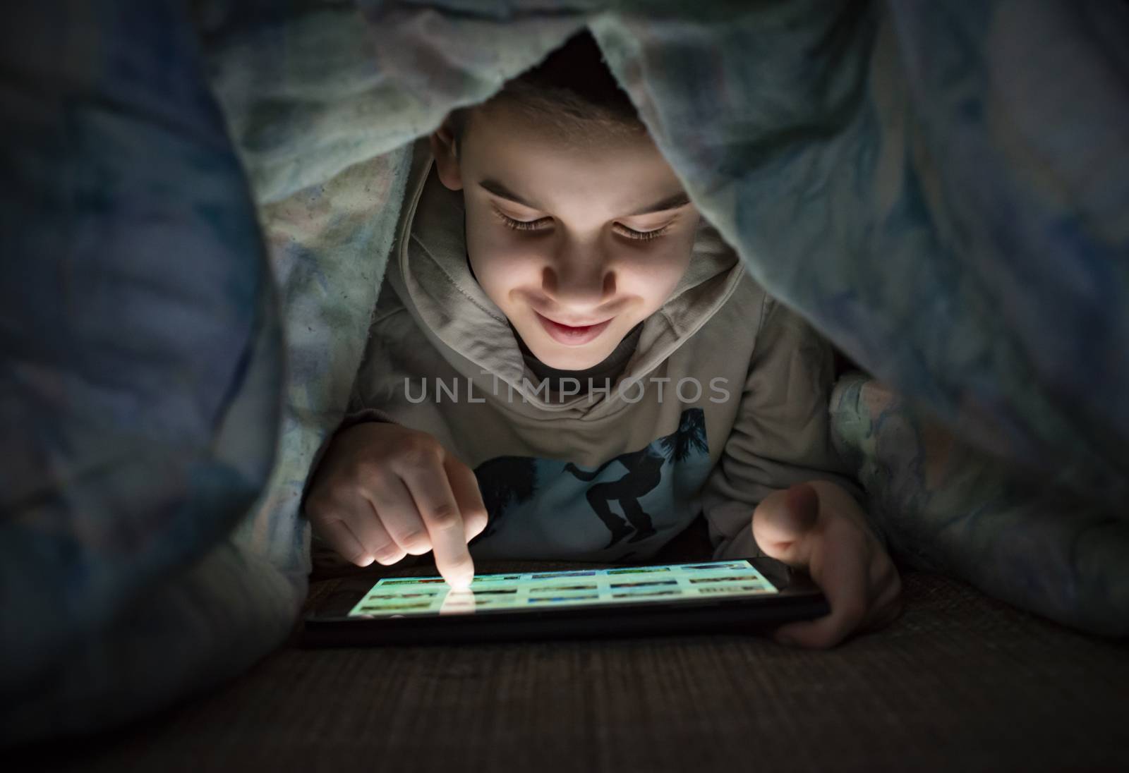 Child watching his tablet in the bed. Illuminated child face from device screen. Smiling Boy under the covers hold a tablet. Night time.