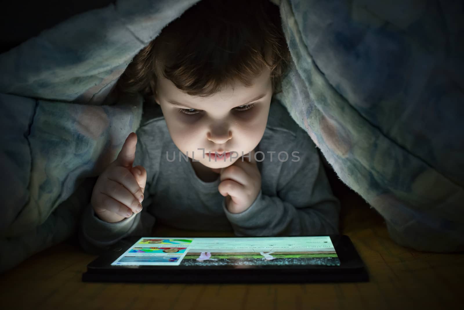 Little girl watching her tablet in the bed. Illuminated child face from device screen. Child dressed with pajamas under the covers hold a tablet. Night time.