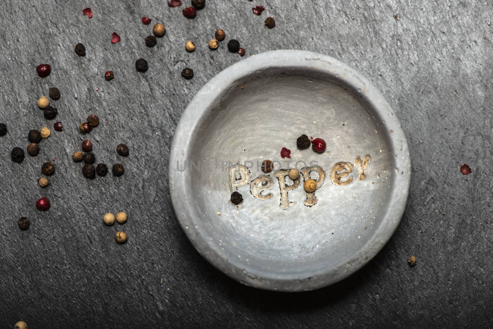 Black pepper in small bowl for spices on dark background. Red, green and black pepper grains close-up and natural light on it. Dark stone background and bowl with text Pepper on the bottom.