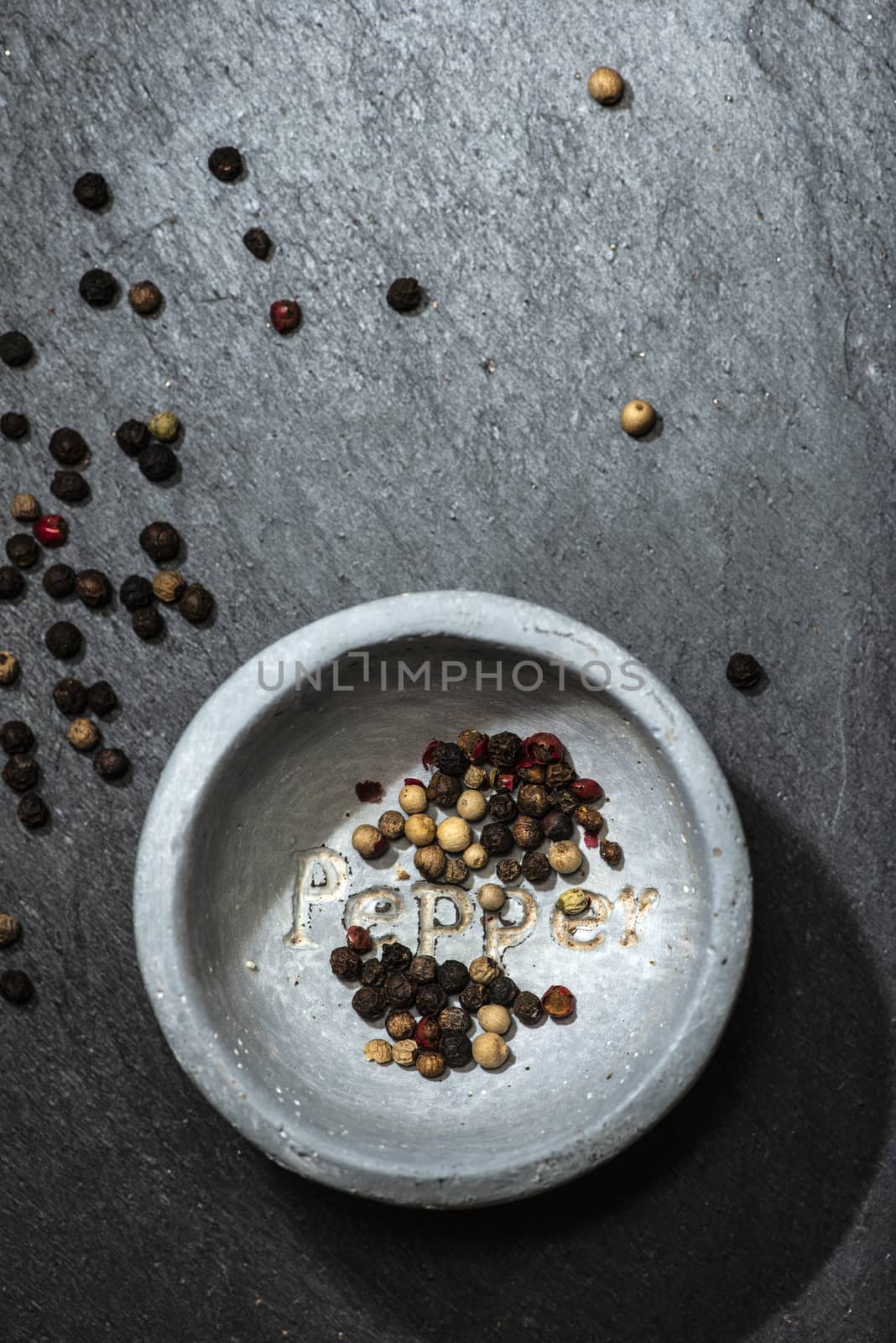 Black pepper in small bowl for spices on dark background. Red, green and black pepper grains close-up and natural light on it. Dark stone background and bowl with text Pepper on the bottom.