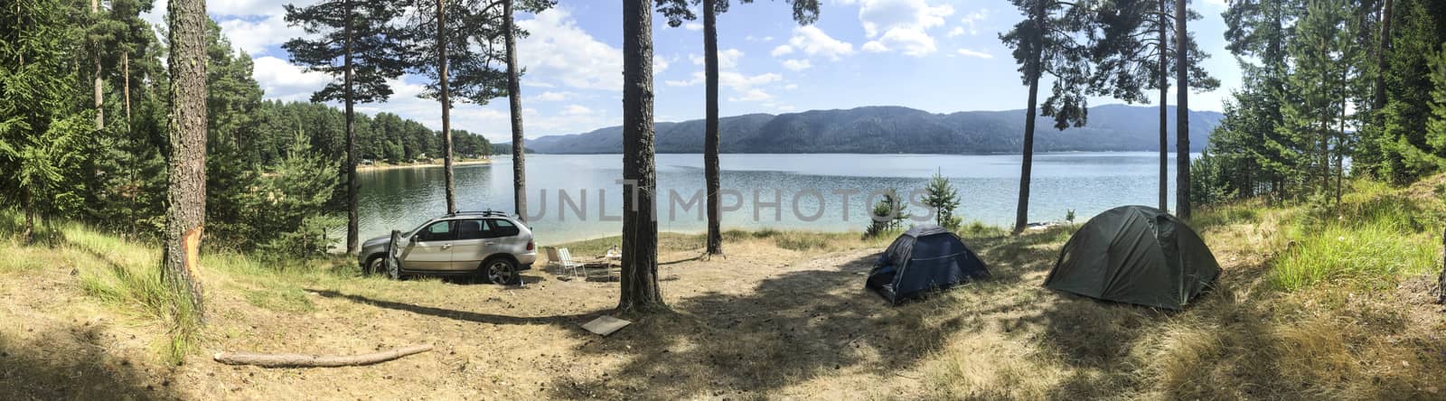Panoramic image of forest and mountain lake. Tents and car in the wild nature. Sunny day. Many pine trees. Panorama in the forest.