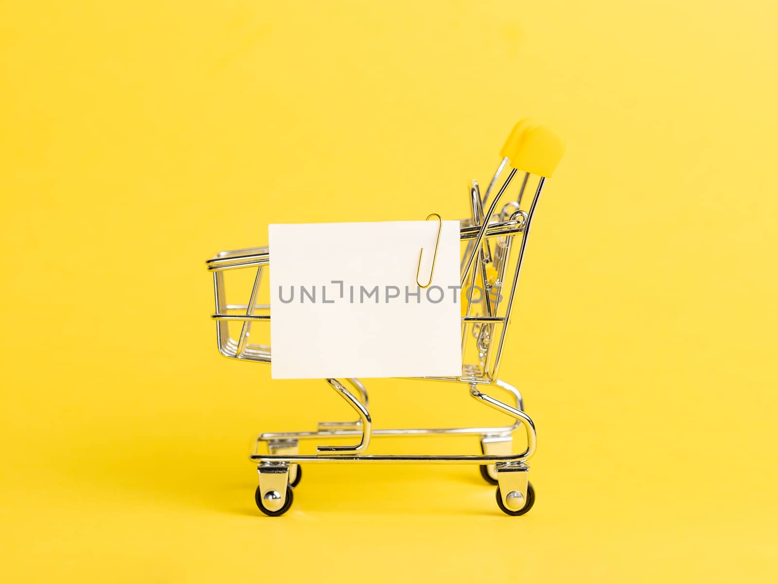 Shopping cart and white paper note list over yellow background. Shopping concept on bright yellow background. Empty white paper note over shopping cart. Copy space for text or design.