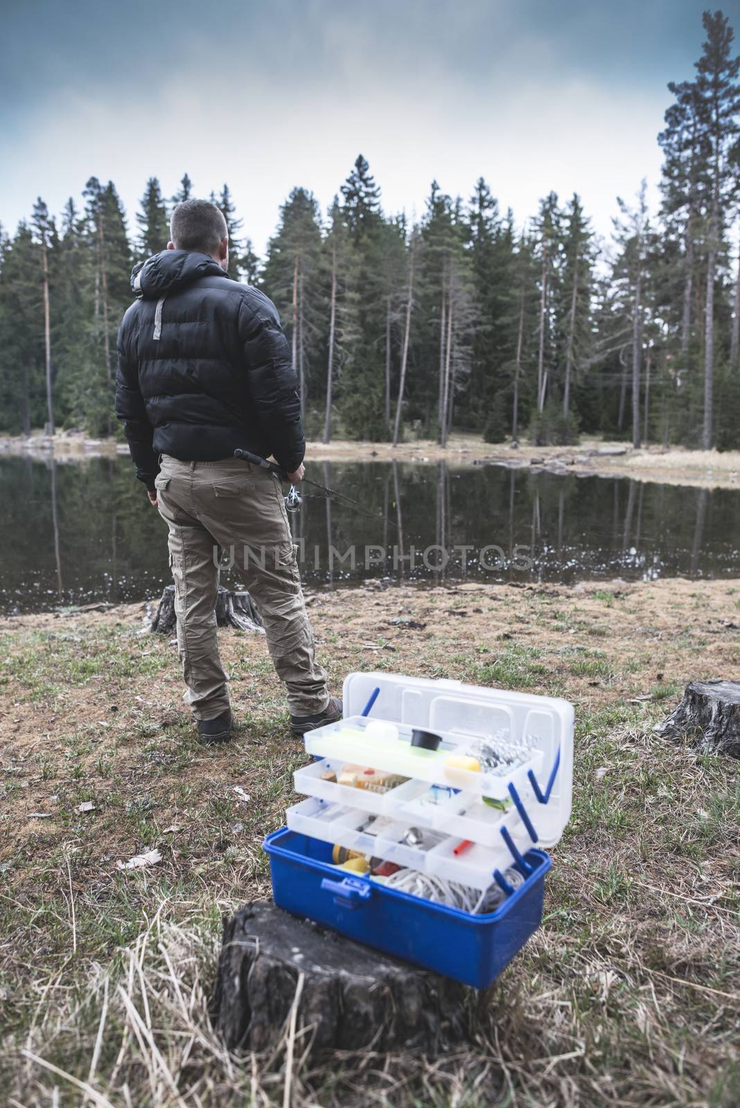 Man fishing on mountain lake. Suitcase with fishing tackle on foreground. Casual clothes