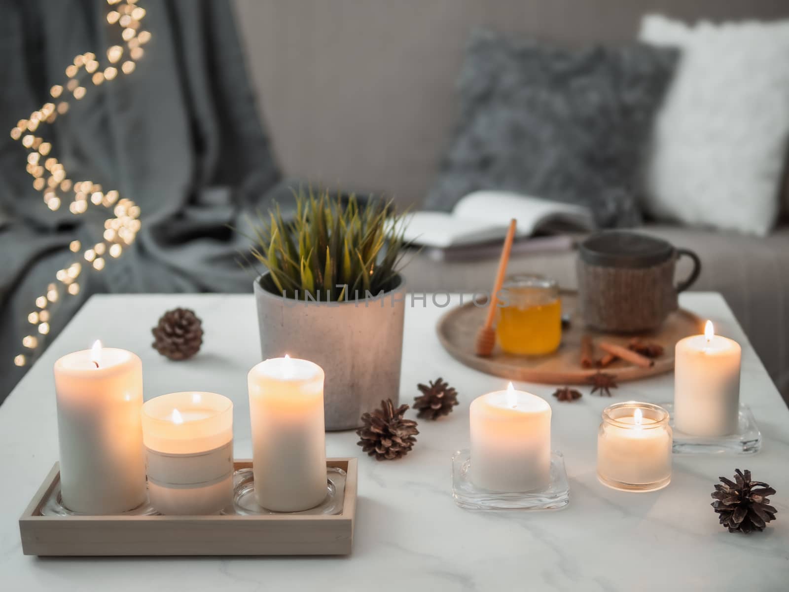 Cozy home, hygge, cosiness concept - burning white fragrance candles on white marble table near sofa with pillows and plaid. Winter decor, hot tea cup and honey